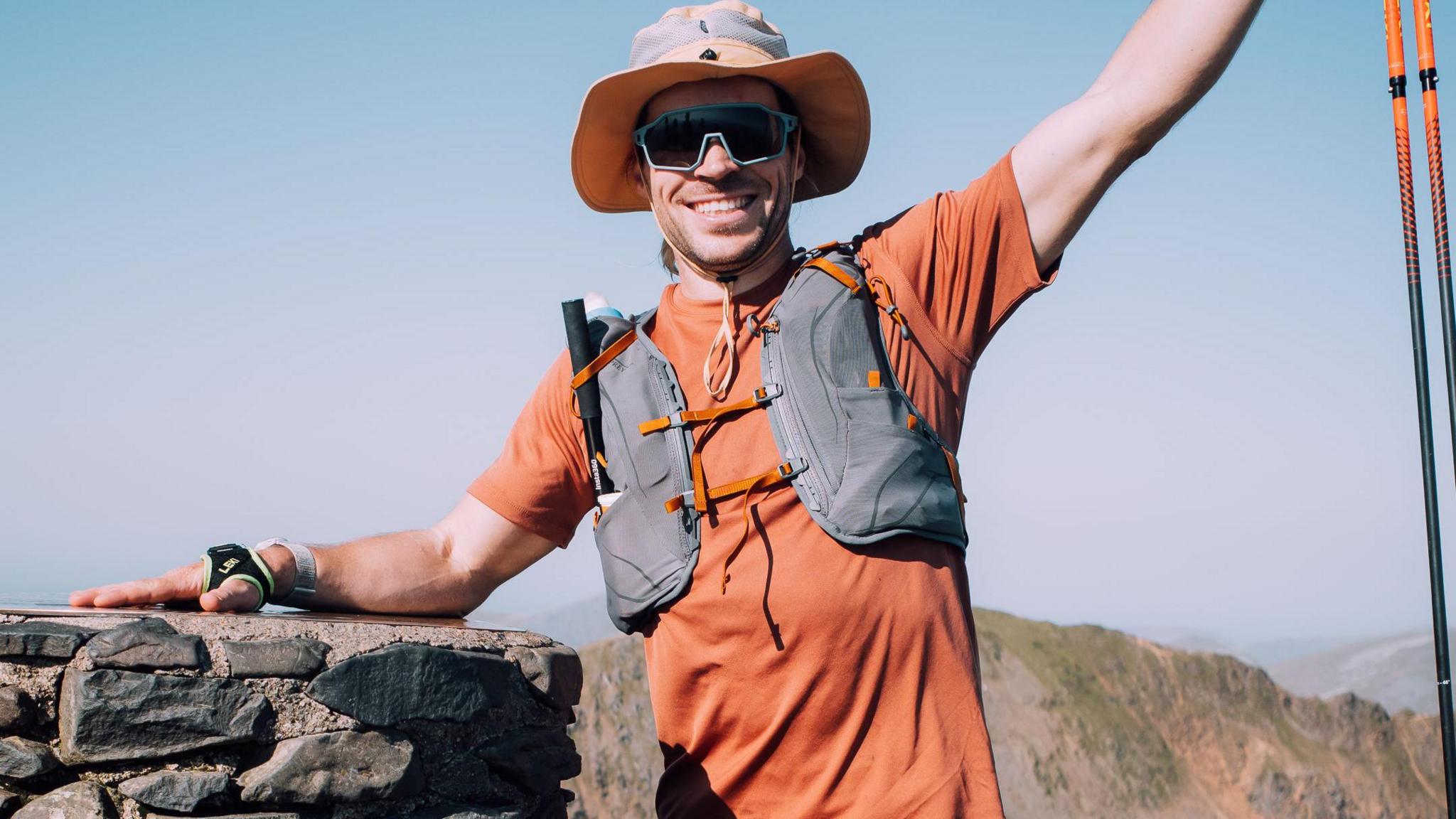 James Norbury pictured on a mountain. He is wearing a hat, glasses and an orange t-shirt. He is equipped with hiking gear, including hiking sticks. He is smiling with his arm raised. 