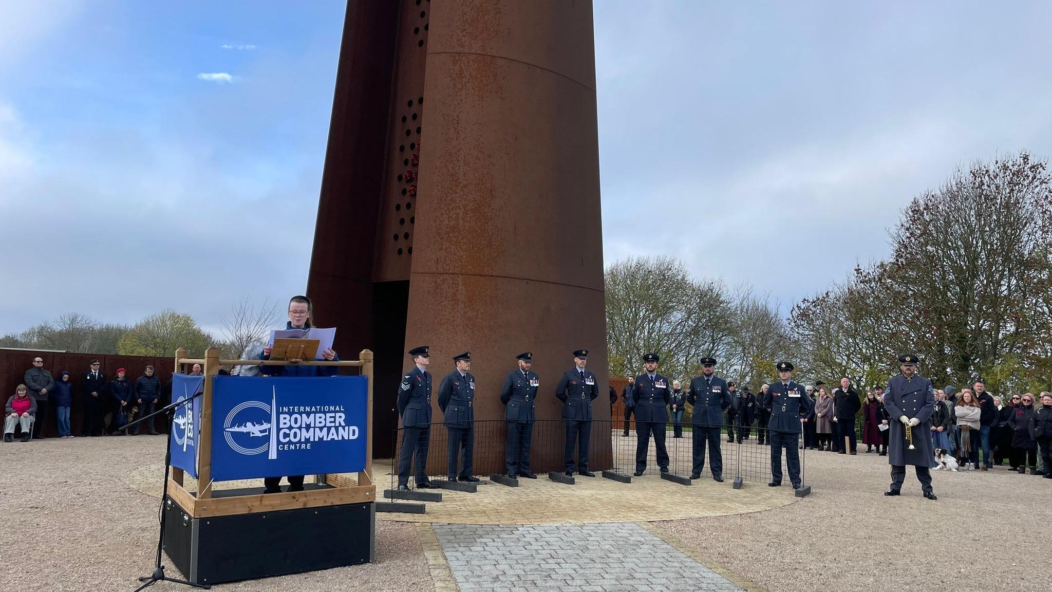 Men in dark navy uniforms and caps stand in a semi circle around the steel memorial. A vicar is standing at a podium reading from a script.
