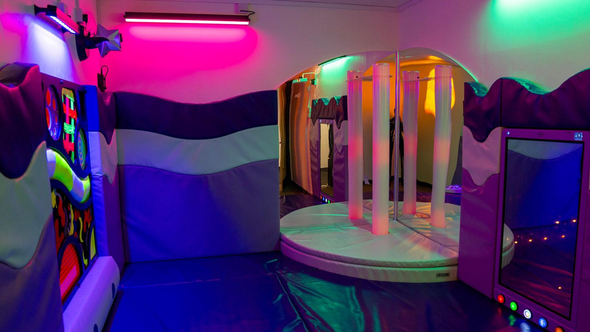 A sensory room which has different colour mats on the floor and on the walls. On the left wall there are different lights with patterns, and to the right there are three mirrors and a small white raised platform. 