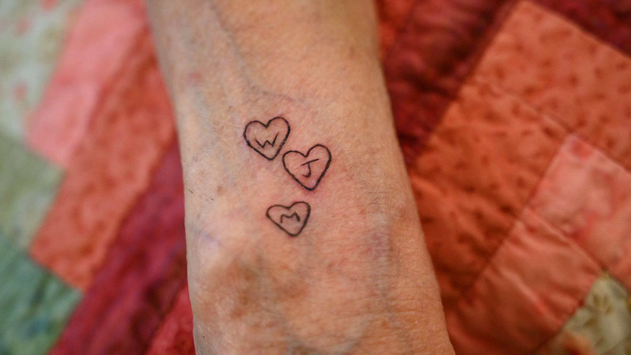 A close-up of an arm with three small hearts tattooed on it. Each heart contains a letter - W, J and M.