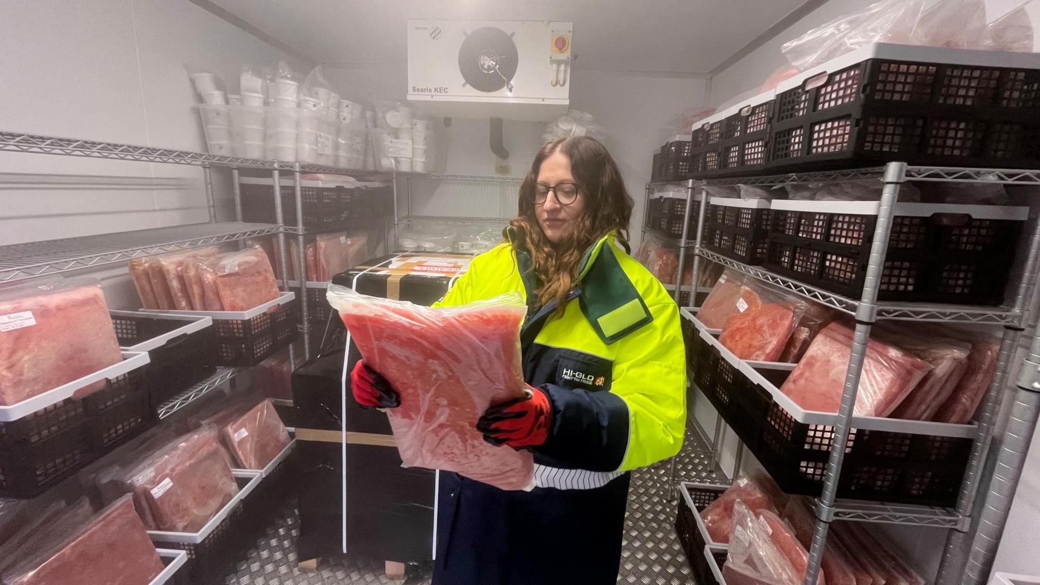 A member of staff from the research institute holds up a packet of frozen tomatoes. They are mushed up inside a packet the size of an A3 folder. The packet is see through. There are about 40 crates filled with packets of frozen tomatoes in a big walk in freezer. The member of staff is wearing a thick bright yellow coat with padded red gloves. It is minus 40 degrees celcius.