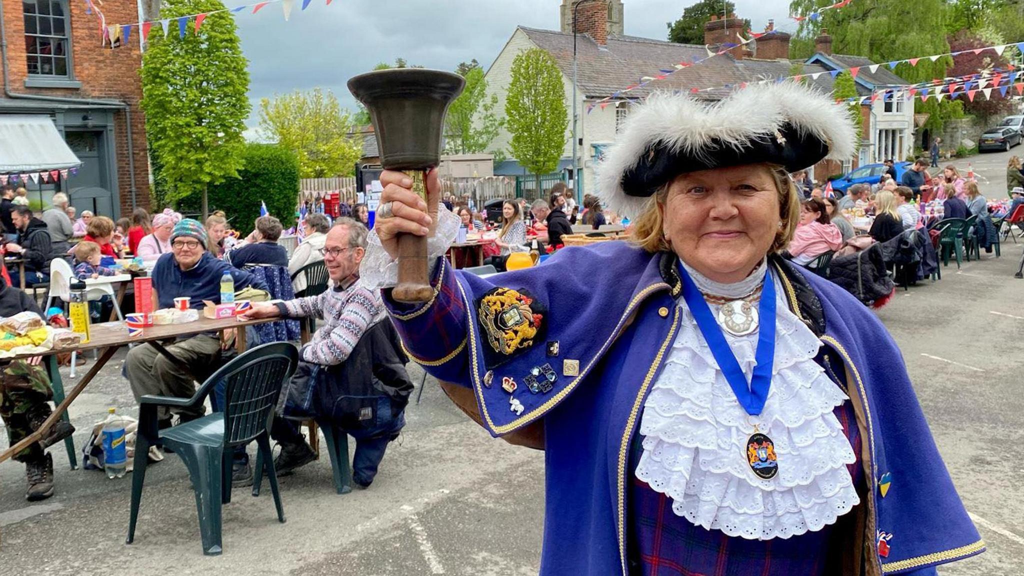 Sue Blower in a blue and white town crier uniform, ringing a bell with bunting and people sat at long tables at an outdoor event behind her