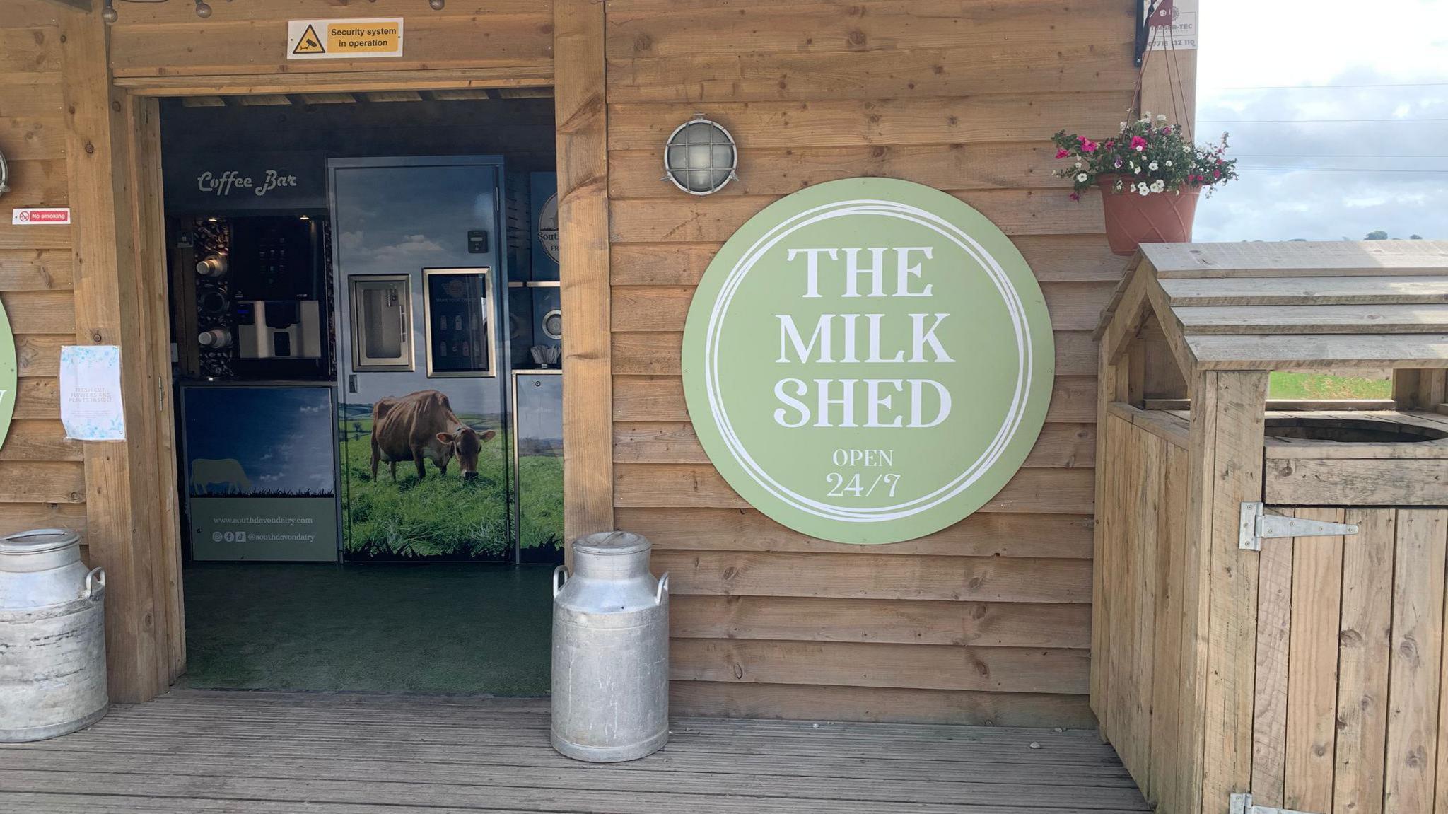 This picture shows a small wooden building labeled "THE MILK SHED", which appears to be a self-service dairy vending facility.
Two metal milk cans are placed at the entrance.
A vending machine area is visible, decorated with images of cows grazing in a field.
A small hanging flower pot is on the right side.
A security system sign is mounted above the entrance.