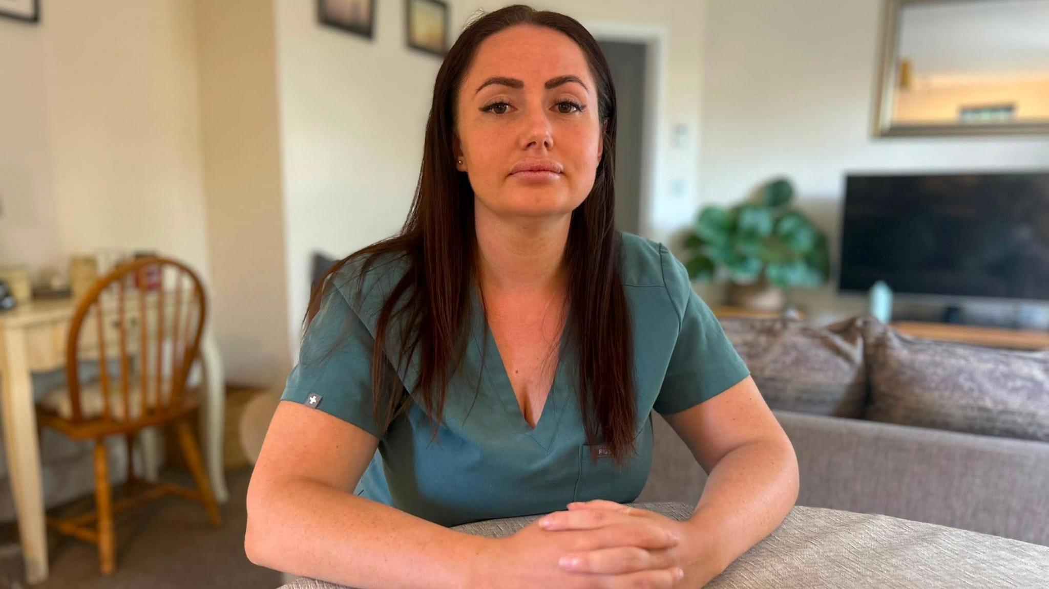 Emily Withington sits at a table in a living room wearing teal dental scrubs, her hands are folded on the tabletop as she faces the camera. The room is bright and includes a sofa, television, houseplant and dining chair in the background.