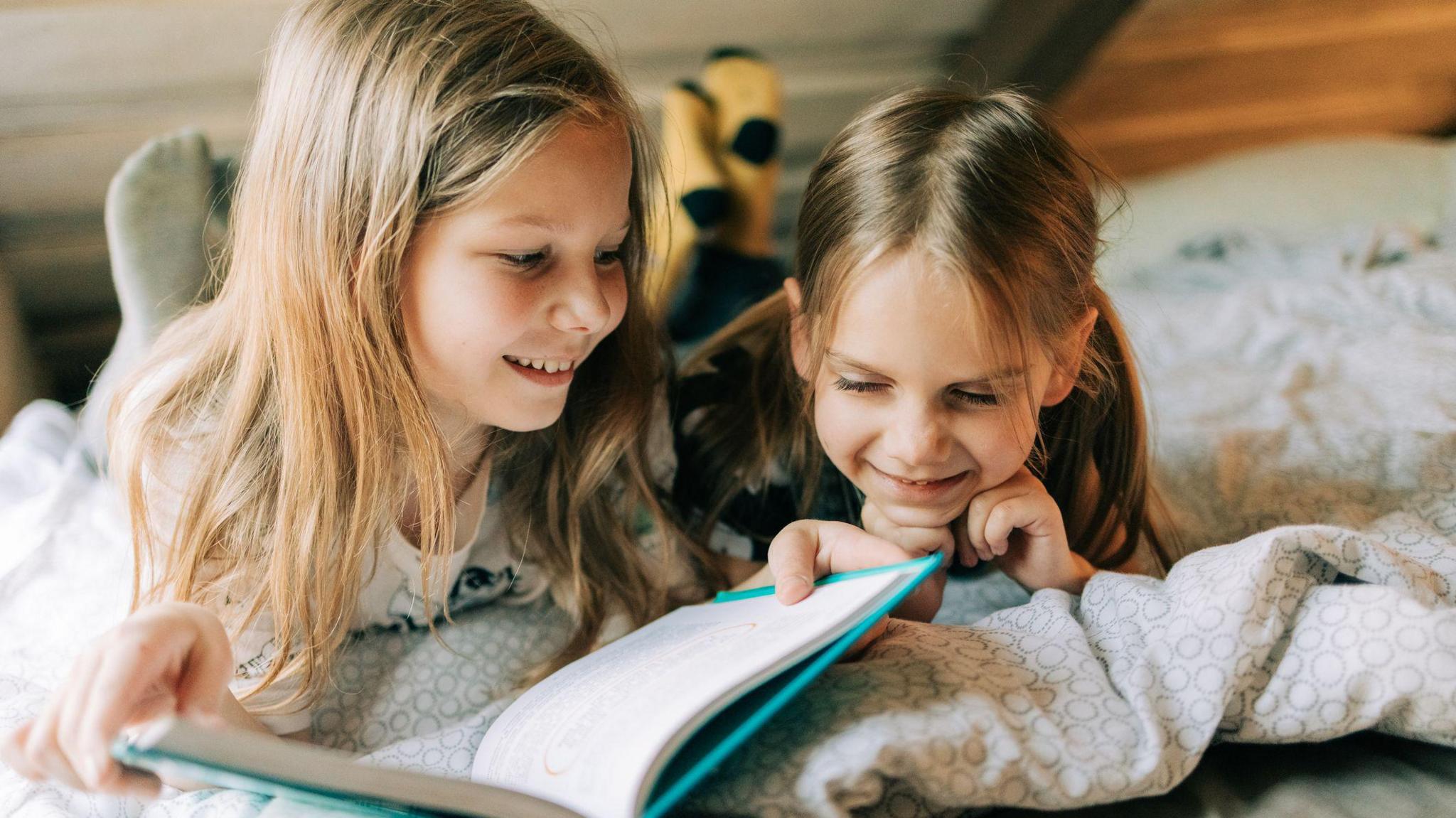 Two girls lie on a bed smiling and reading a book between them.
