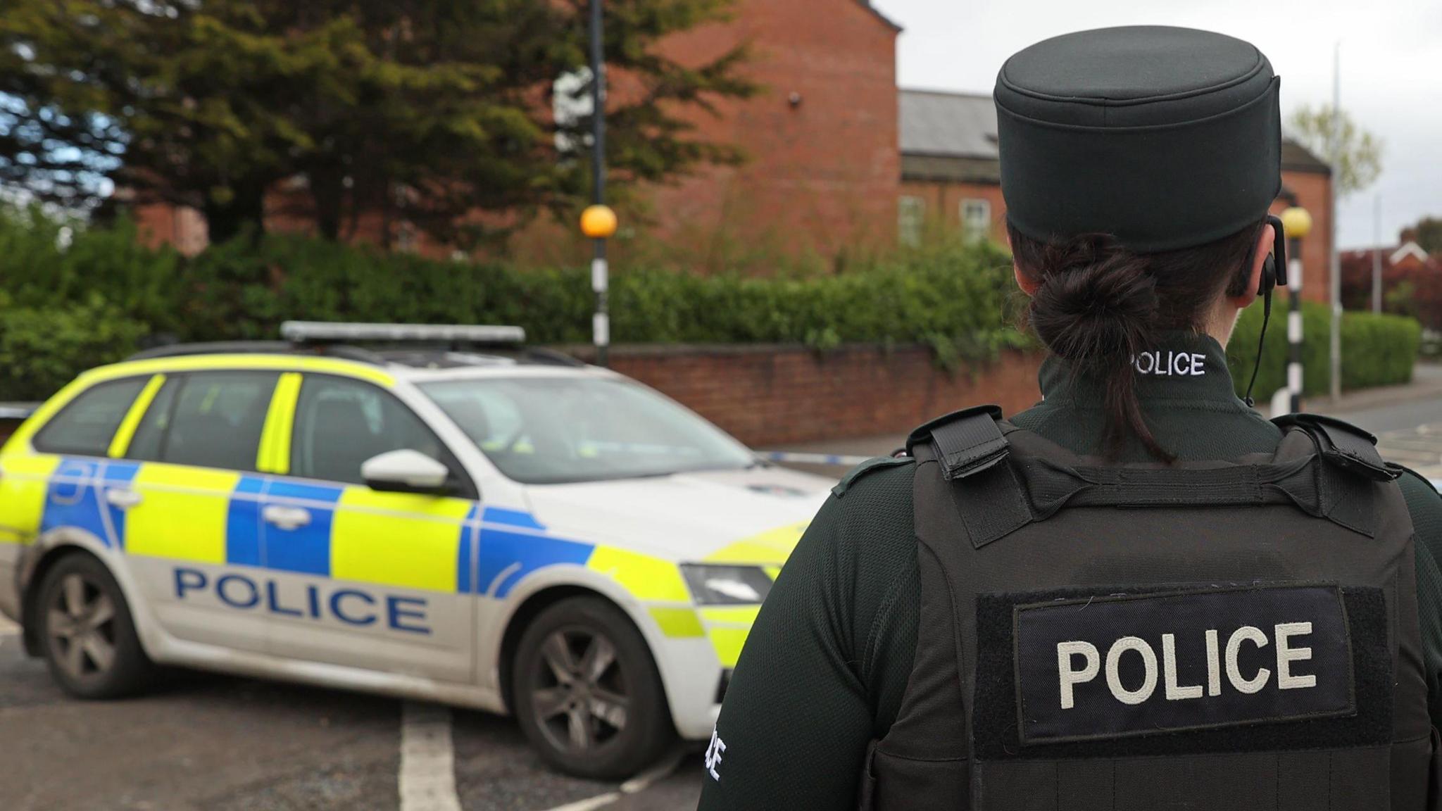 A police officer standing with their back to the camera. The officer has dark brown hair in a bun and is wearing the official uniform with a black bullet proof vest which has "police" written on it. To the left of the officer is a blue, yellow and white police car.