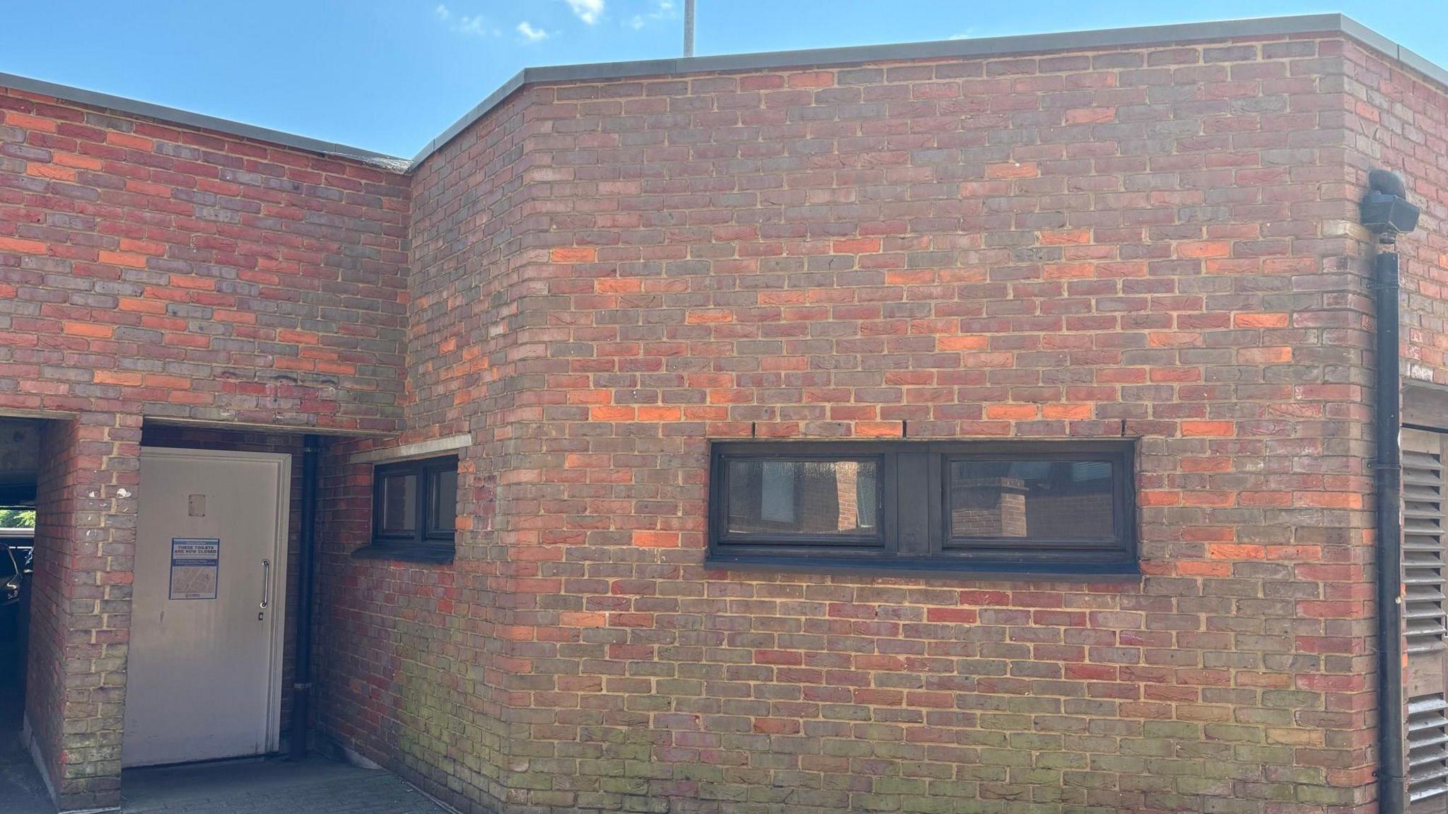 Brick toilet block with a white door, with a sign on it, windows and a down pipe.