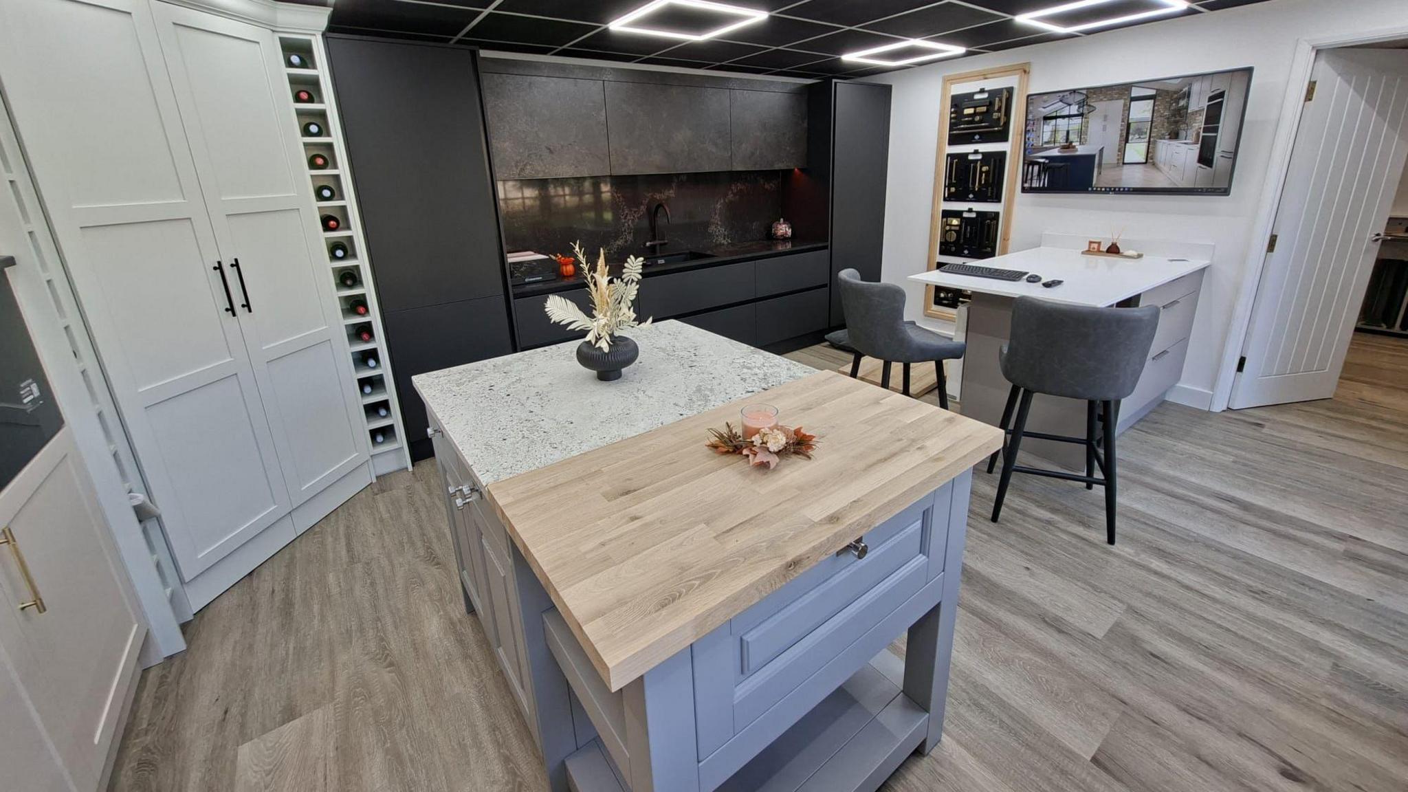 This is a picture of part of the inside of a kitchen showroom. To the left of the picture is a double door white floor to ceiling cupboard with a wine rack beside it. In the foreground of the picture is a kitchen island with a vase of flowers standing on the top. To the back of the picture are dark grey kitchen units. To the right of the picture there is another kitchen island with two chairs. There are pictures and a mirror on the wall. The ceiling is made of black ceiling tiles with three square LED lights visible.