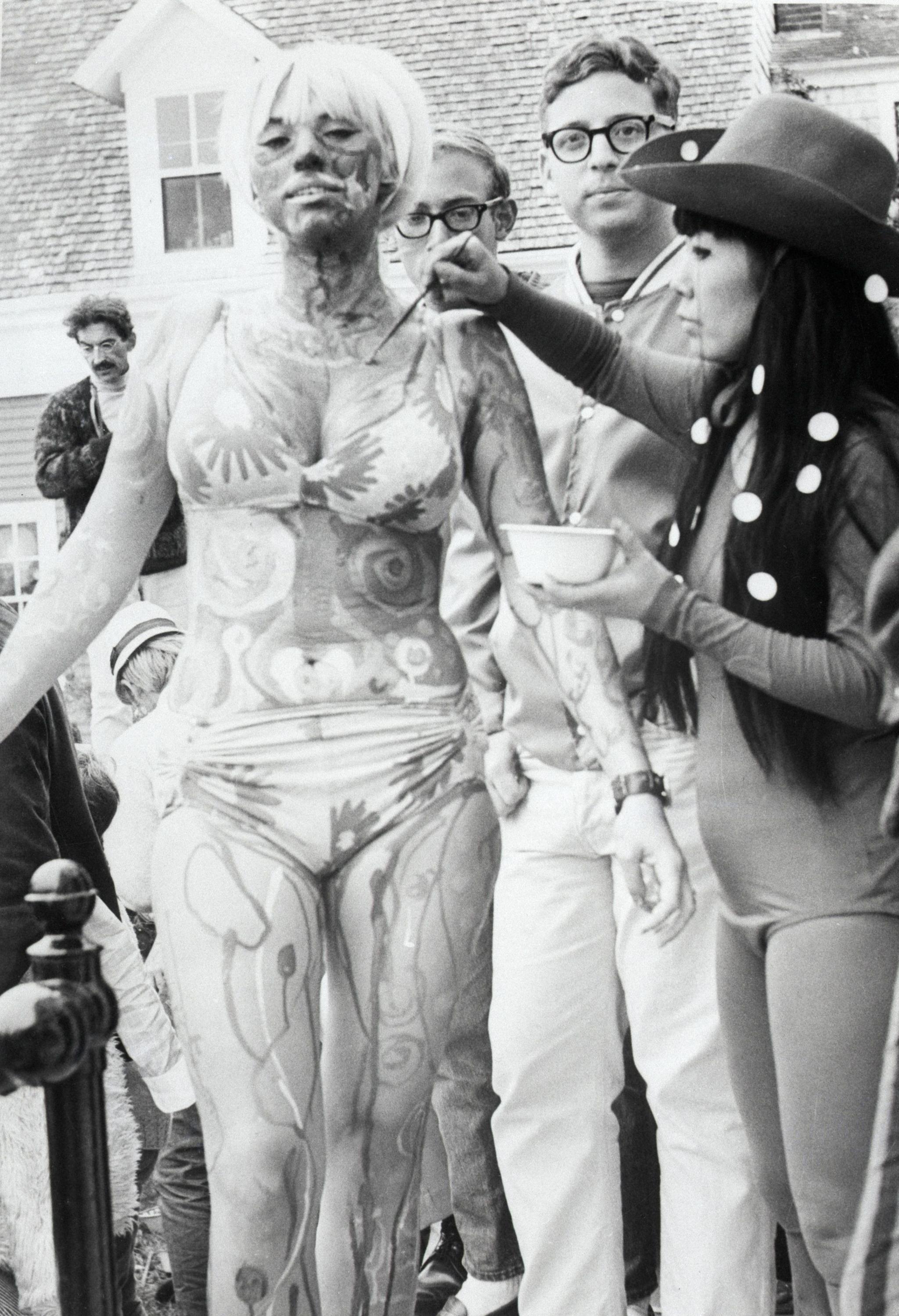 Black & white photo of Yayoi Kusama in 1967 painting a young woman wearing a bikini at a Body Festival in Provincetown, Massachusetts.