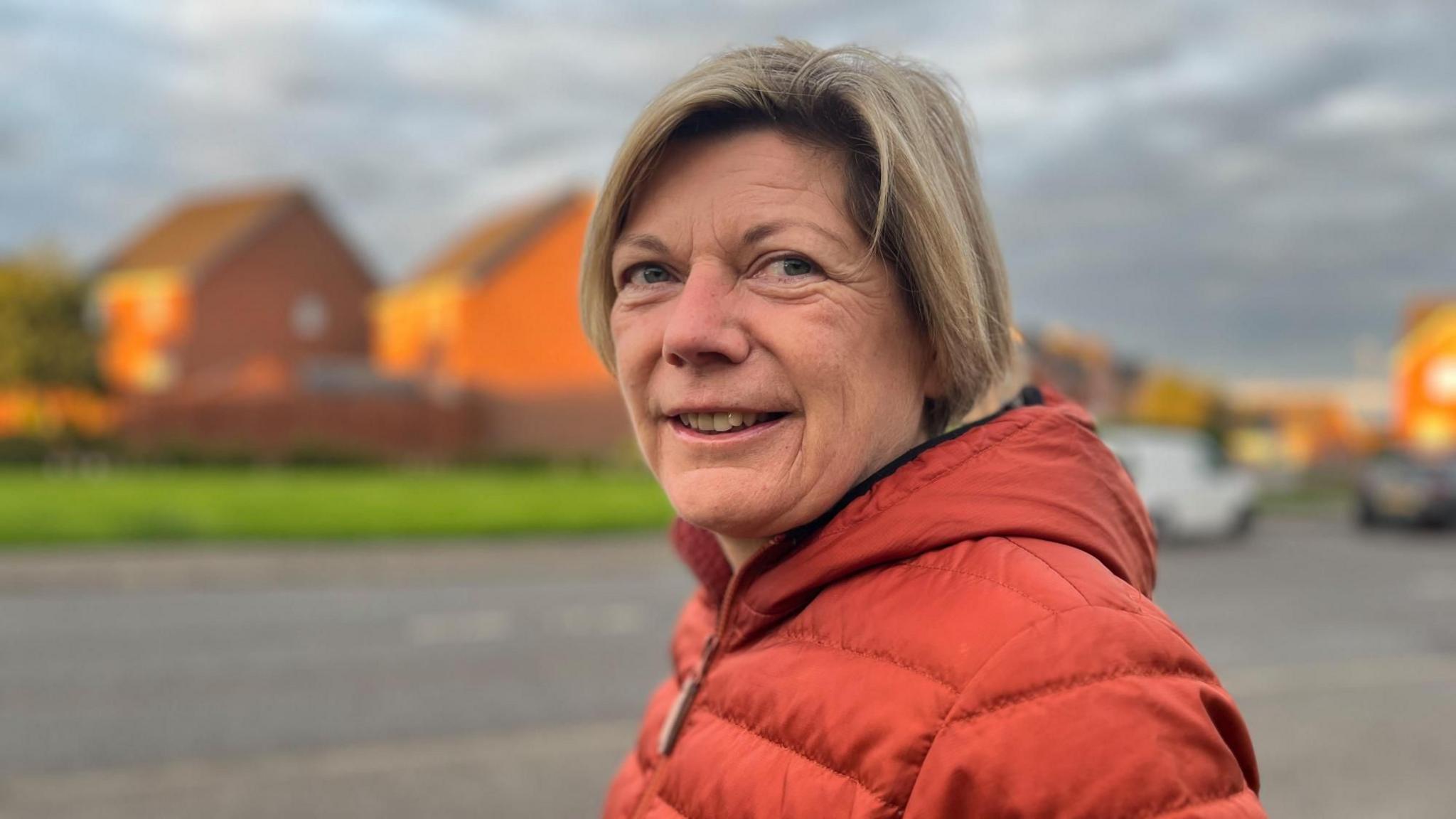 Barbra Clegg standing outside next to a road with houses in the background. She has short, blonde hair and is wearing an orange puffer jacket