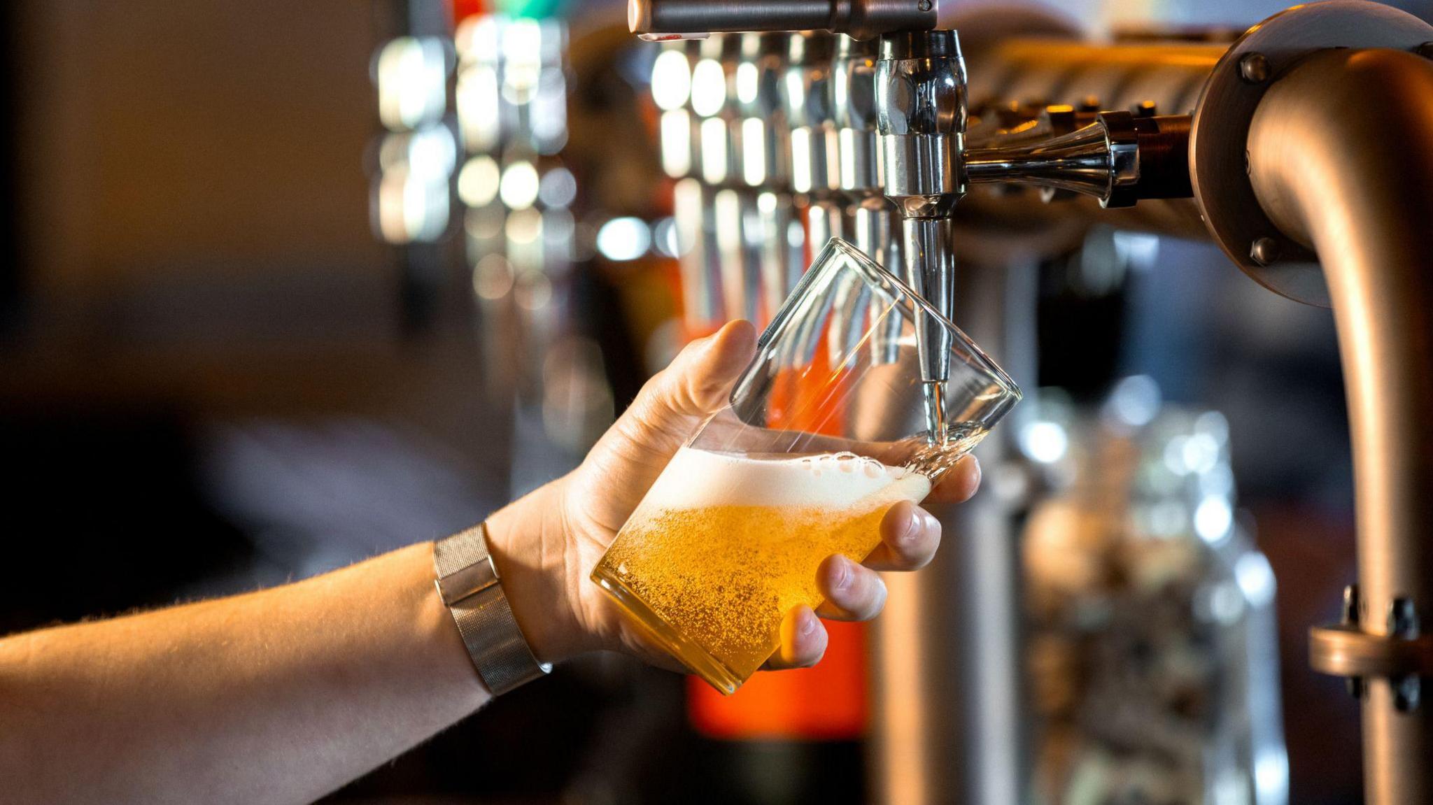 A close-up of an unrecognisable person's hand holding a pint glass at an angle being filled with beer. The beer is golden in colour, creating foam and bubbles.
