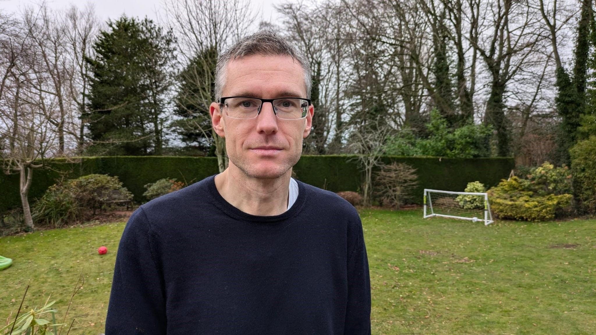 Mark Church is standing in a garden with a hedge behind him. He is a man in his 40s with glasses and wearing a blue jumper.