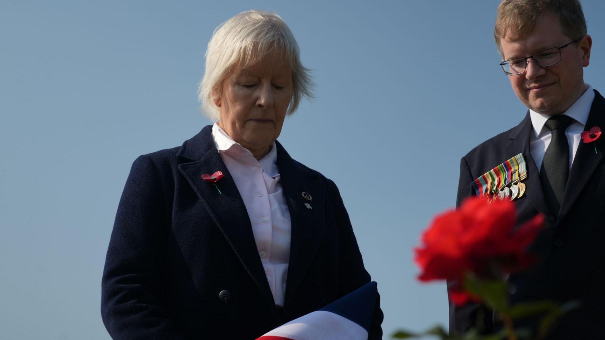 Major Angier’s daughter, Tabby, by her father's grave; a woman in a dark jacket looks down, a man stands to her right.