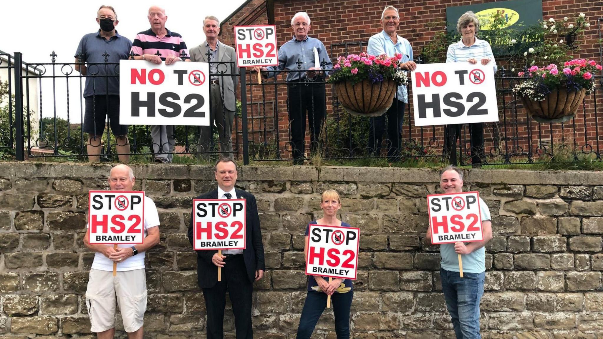 A group of people stand holding placards with the words, "STOP HS2" written on. Some are standing with their backs to a stone wall and others stand above the wall