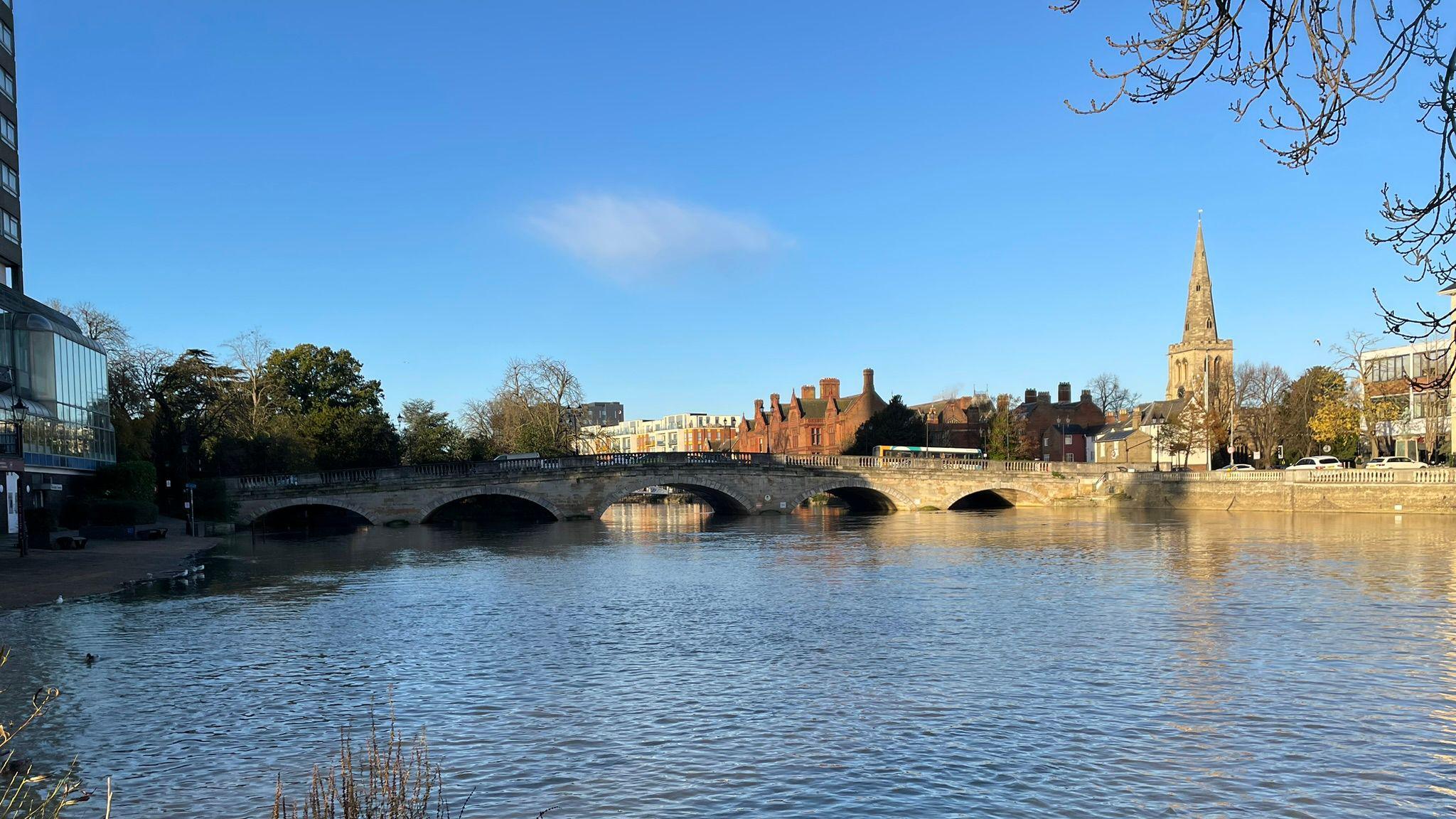 Bedford, showing the river, the Town Bridge and a church to the left. The river level is very high. There are buildings in the distance and the sky is blue. 