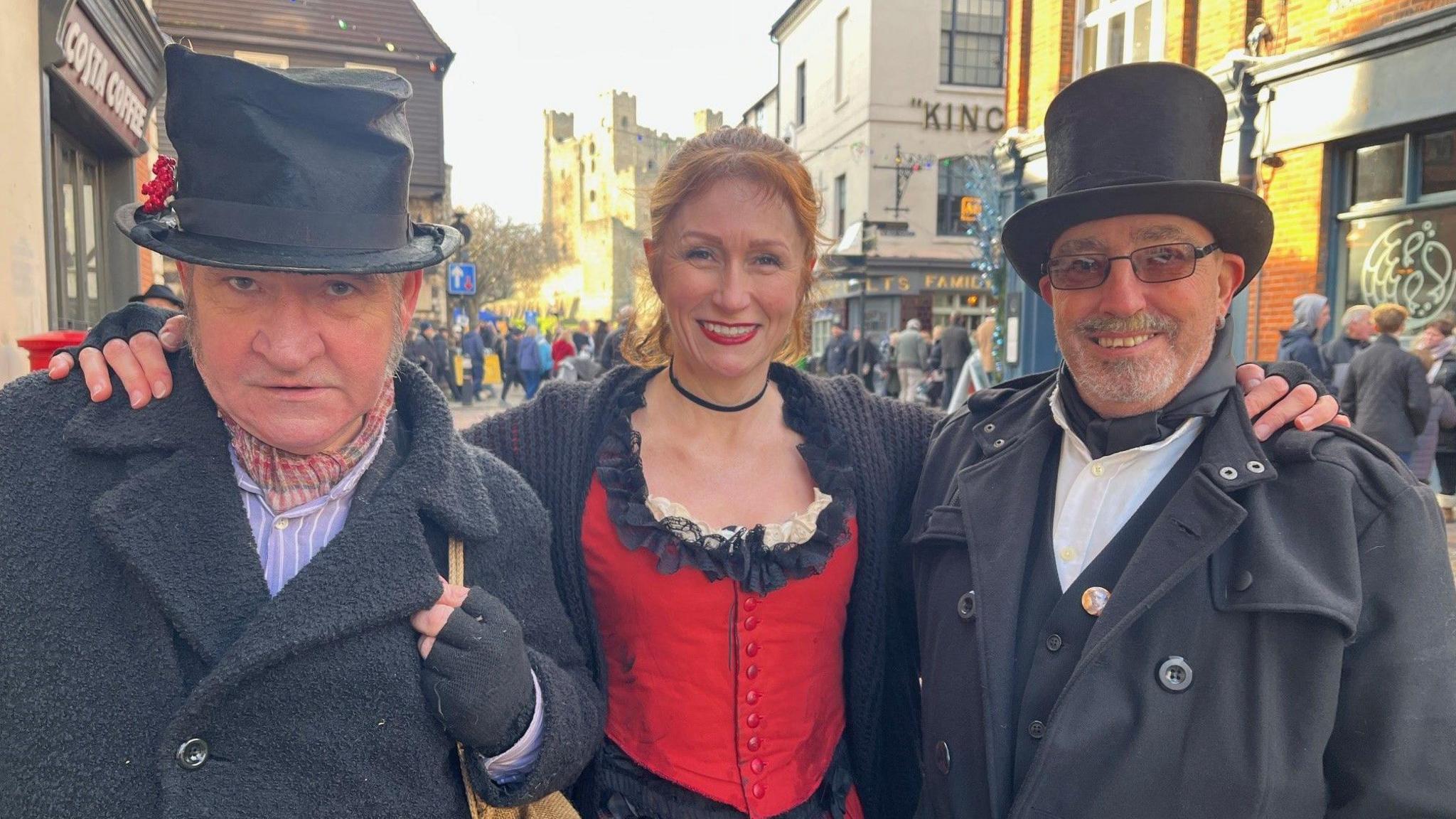 Three people dressed in Victorian inspired costume - two in black suits and top hats and one in a red dress.