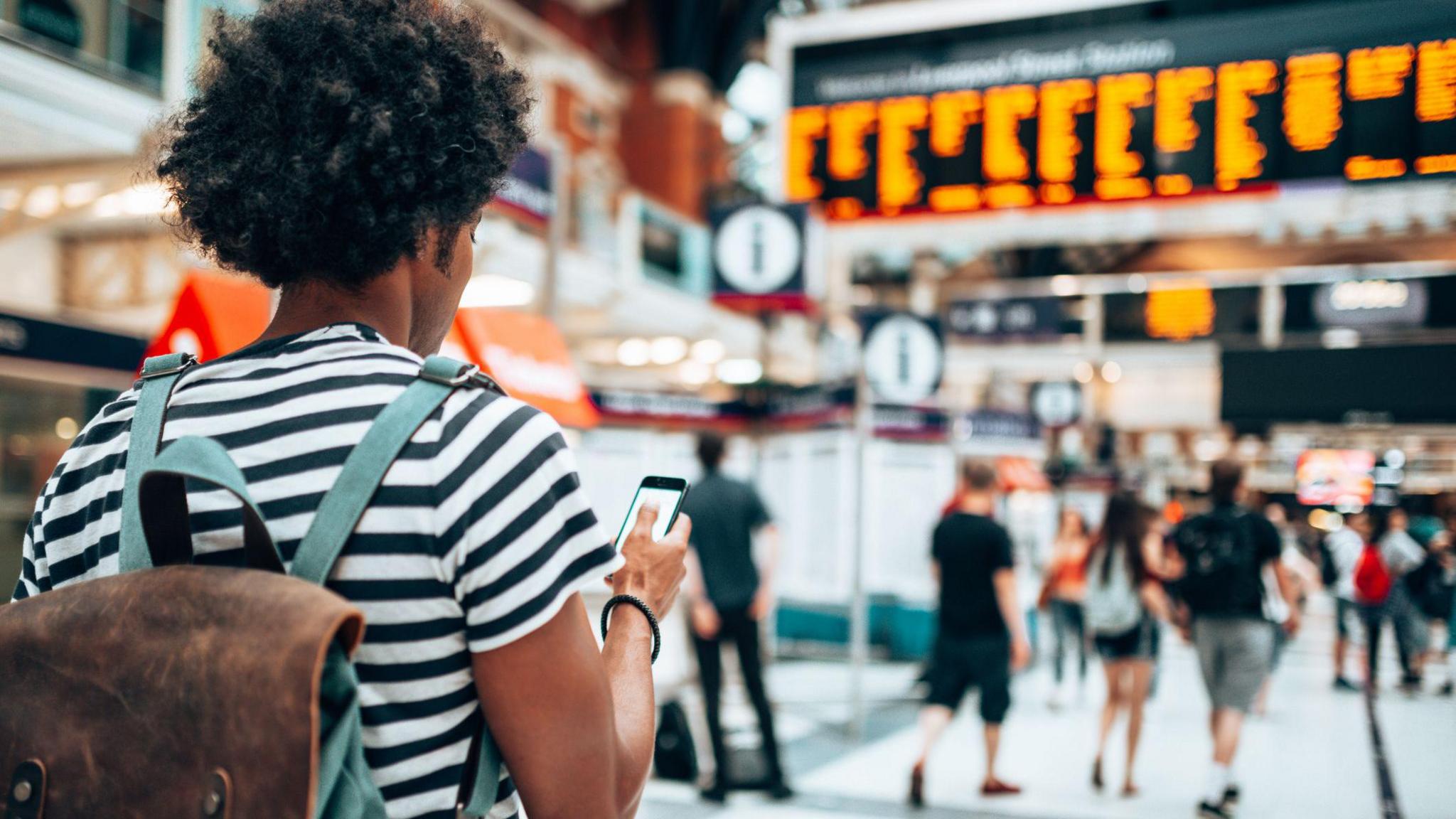 Commuter stands with phone in hand at a busy railway station.