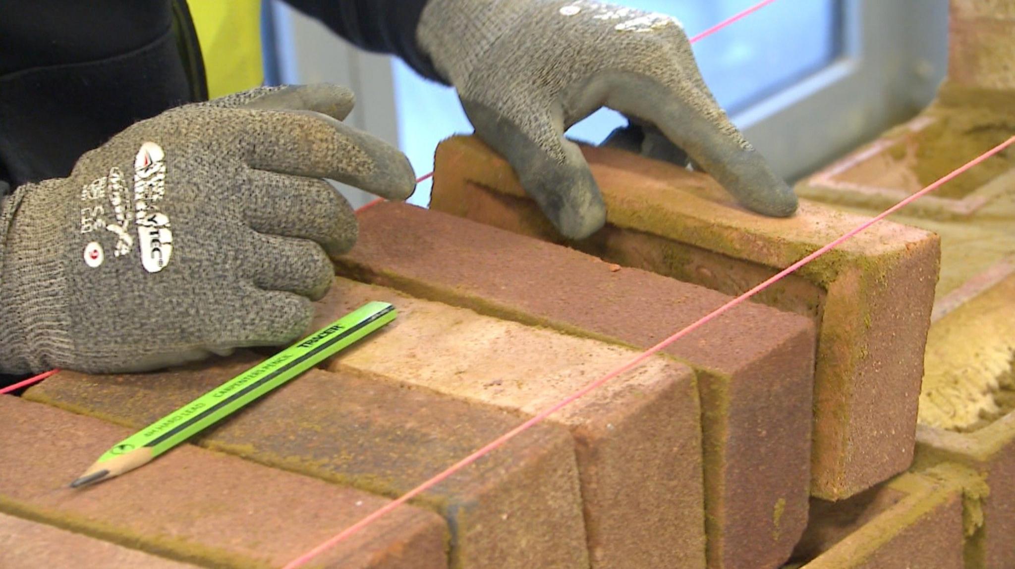 A close-up image of gloved hands with one holding a brick and the other resting on a row of bricks. There is a pencil and red string in the foreground.