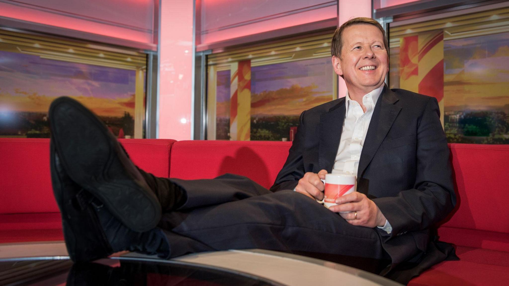 Bill Turnbull sat on the famous red BBC Breakfast sofa while smiling. He has his feet on the table in front of him and is wearing a black suit with a white shirt underneath. Turnbull has short brown hair and is looking away from the camera.