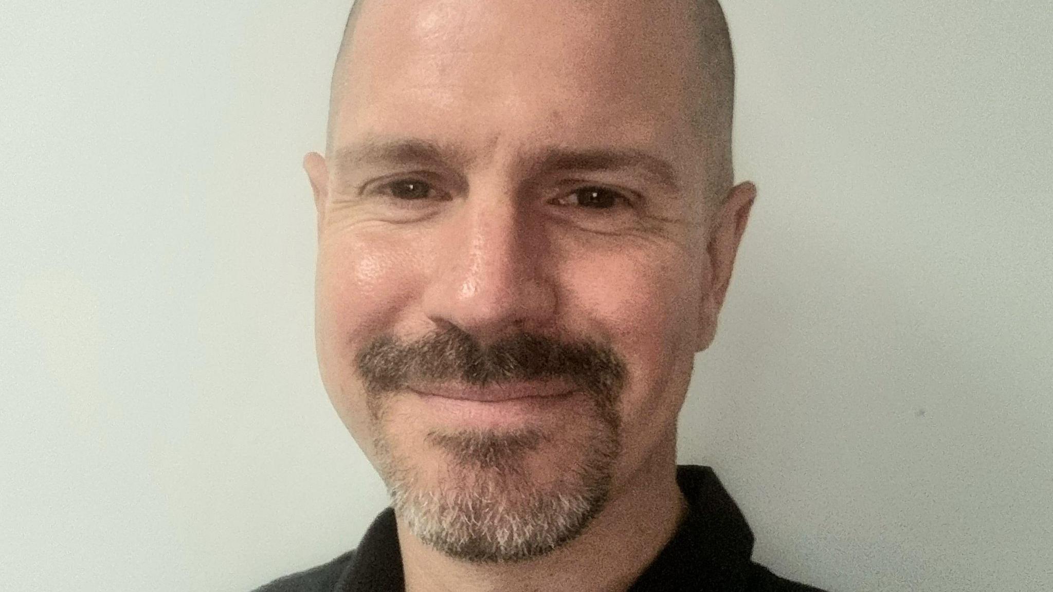 Ross Ayres has brown eyes, a shaven head, moustache and cropped goatee beard. He is wearing a black top.