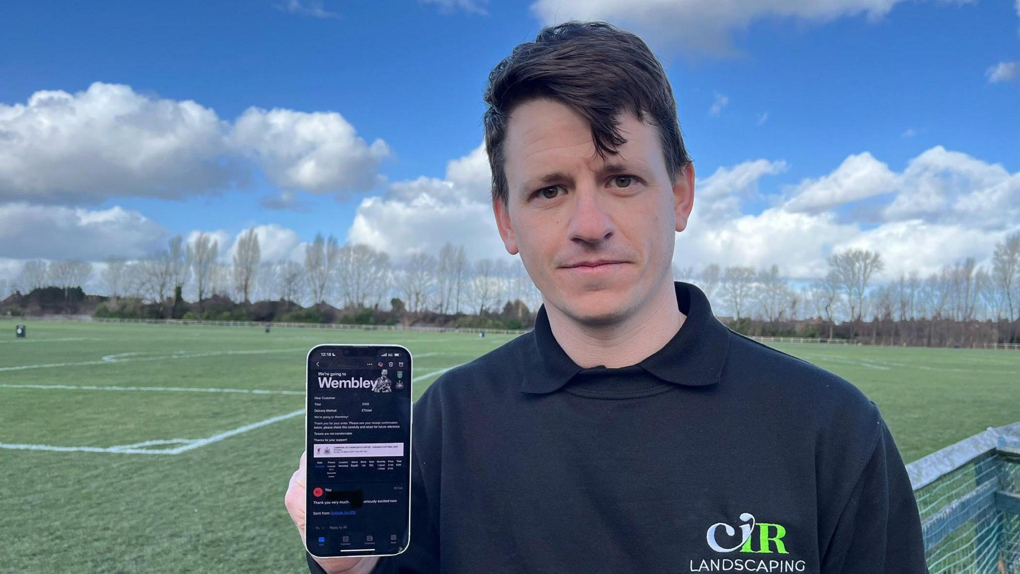 Reece Calvert stands in front of a football pitch holding up his mobile phone. The phone displays a photoshopped email which he was sent by a ticket scammer. 