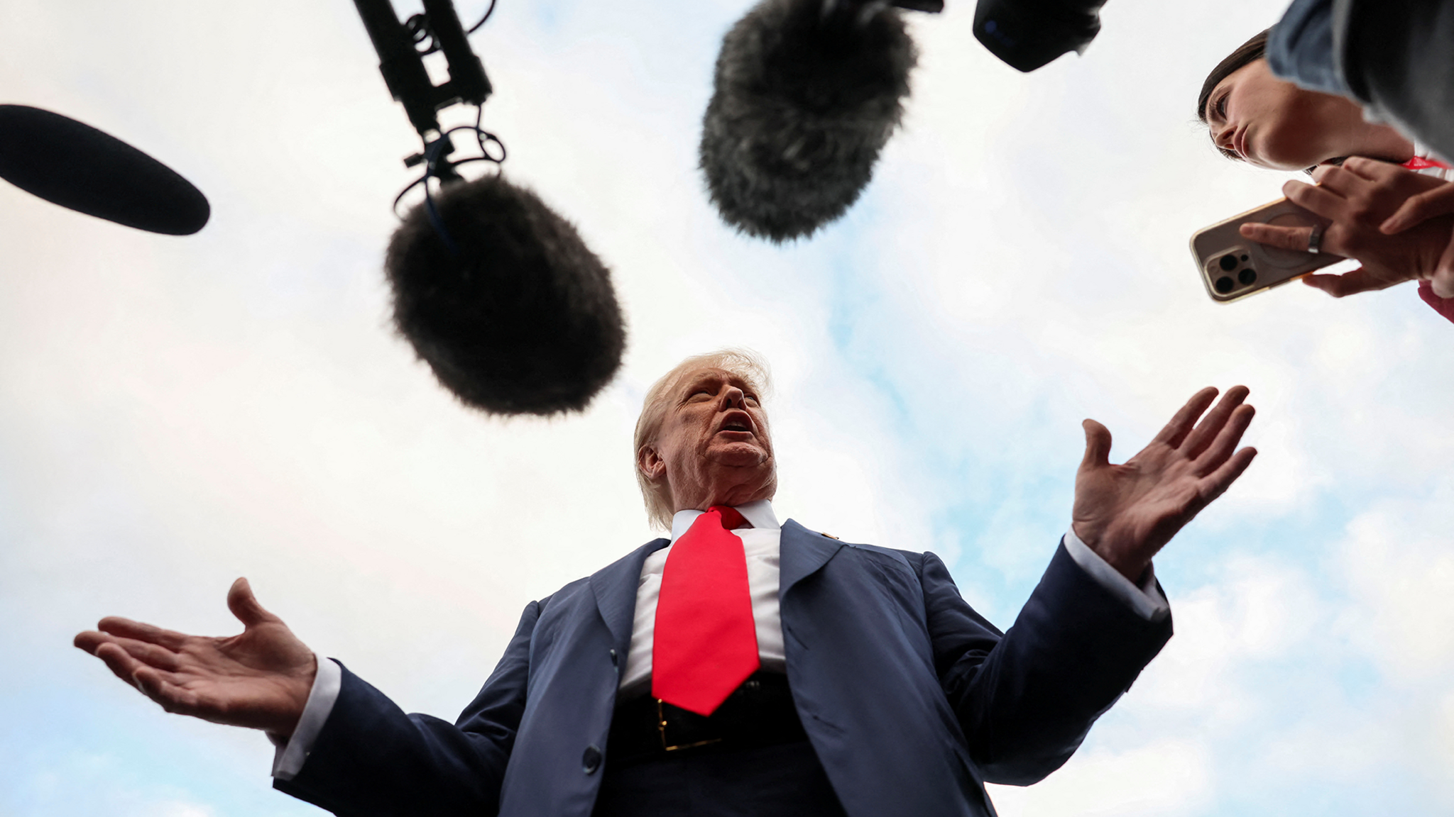 A picture taken from below as President Donald Trump speaks to the media after arriving at Prestwick Airport. He is standing with his arms apart, wearing a blue suit, white shirt and a red tie.