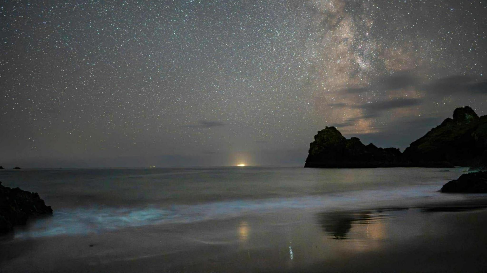 A picture of the view across Cornwall. In the background the sky full of stars can be seen with a body of water and shadow of cliffs surrounding it. At the bottom of the image a strong blue glow can be seen.