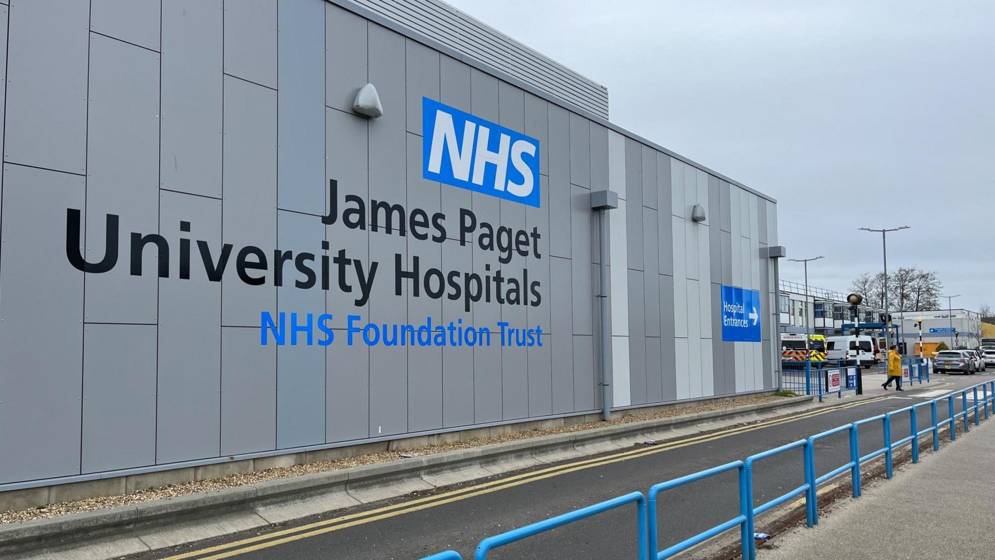 The side of the James Paget Hospital building, in large lettering it says "NHS James Paget University Hospitals NHS Foundation Trust". On the right are ambulances.