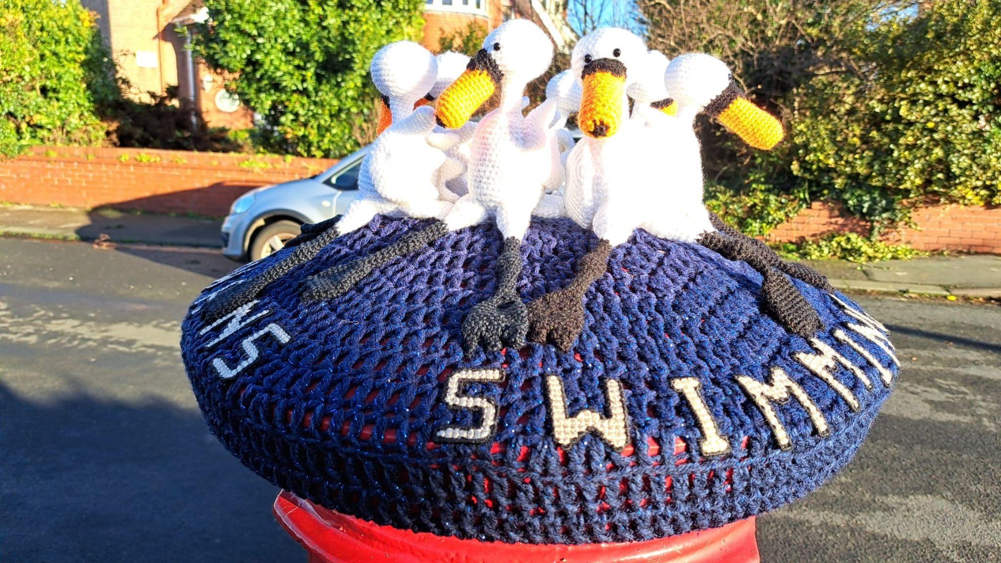 Knitted post box topper showing "seven swans a swimming"