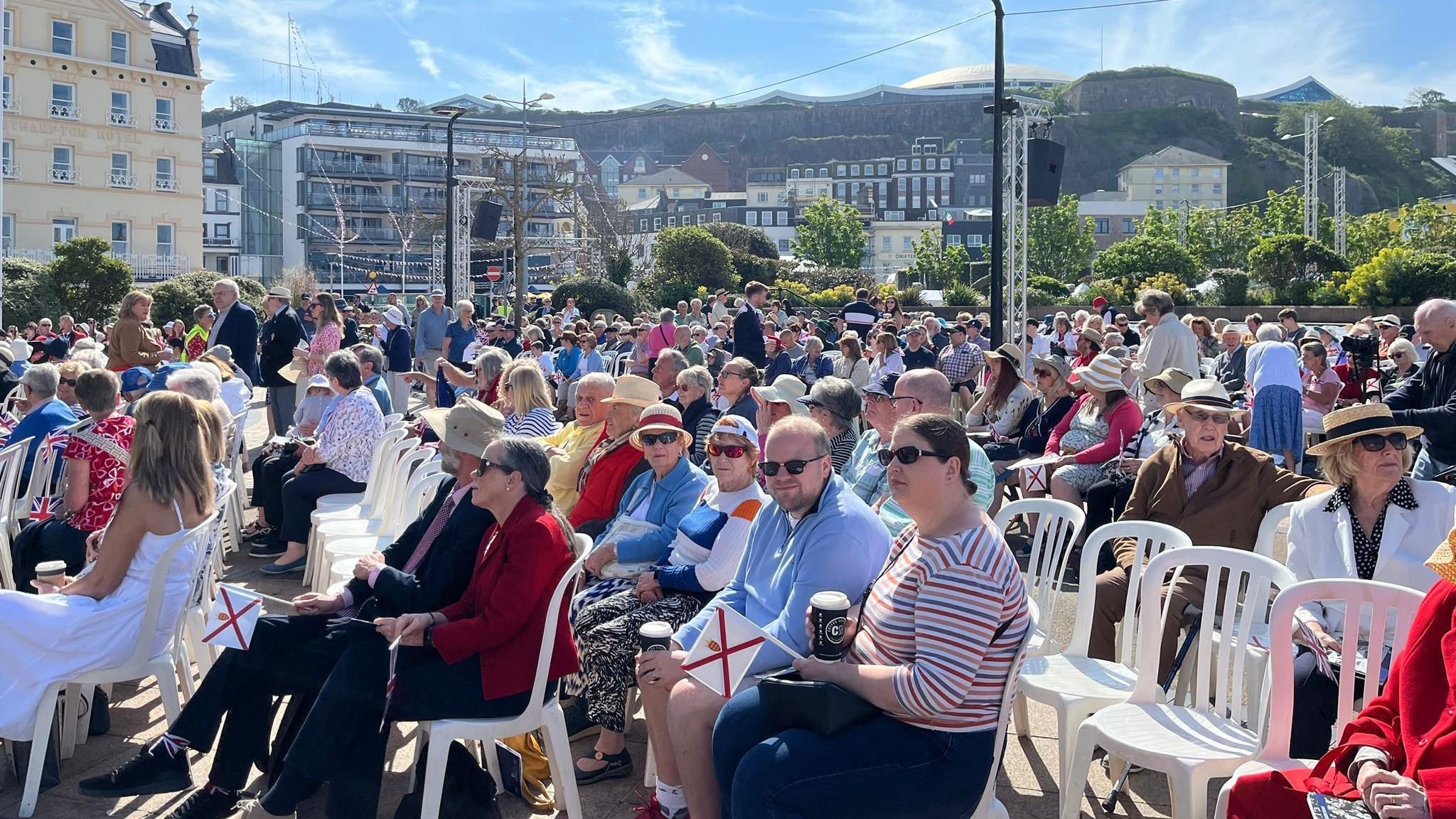 Crowds gather at Liberation Square for Liberation Day Jersey 2024. The sun is shining and there are blue skies. Hundreds of people are sat on chairs as they take part in the celebrations. Islanders and visitors are wearing hats and sunglasses. Some are holding the Jersey flag, which is white with a red cross surmounted by a yellow "Plantagenet crown".