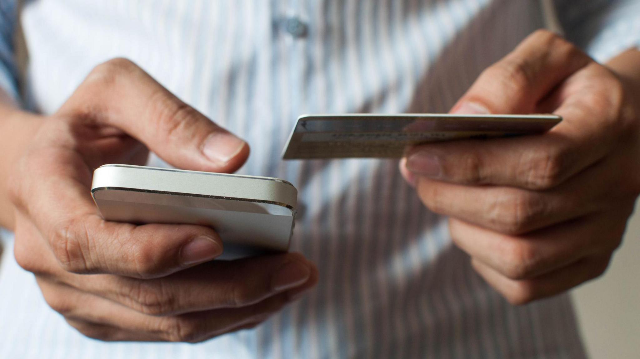 Stock close-up image of a man holding a phone in his right hand and a bank card in the other. He is wearing a blue-and-white striped shirt.