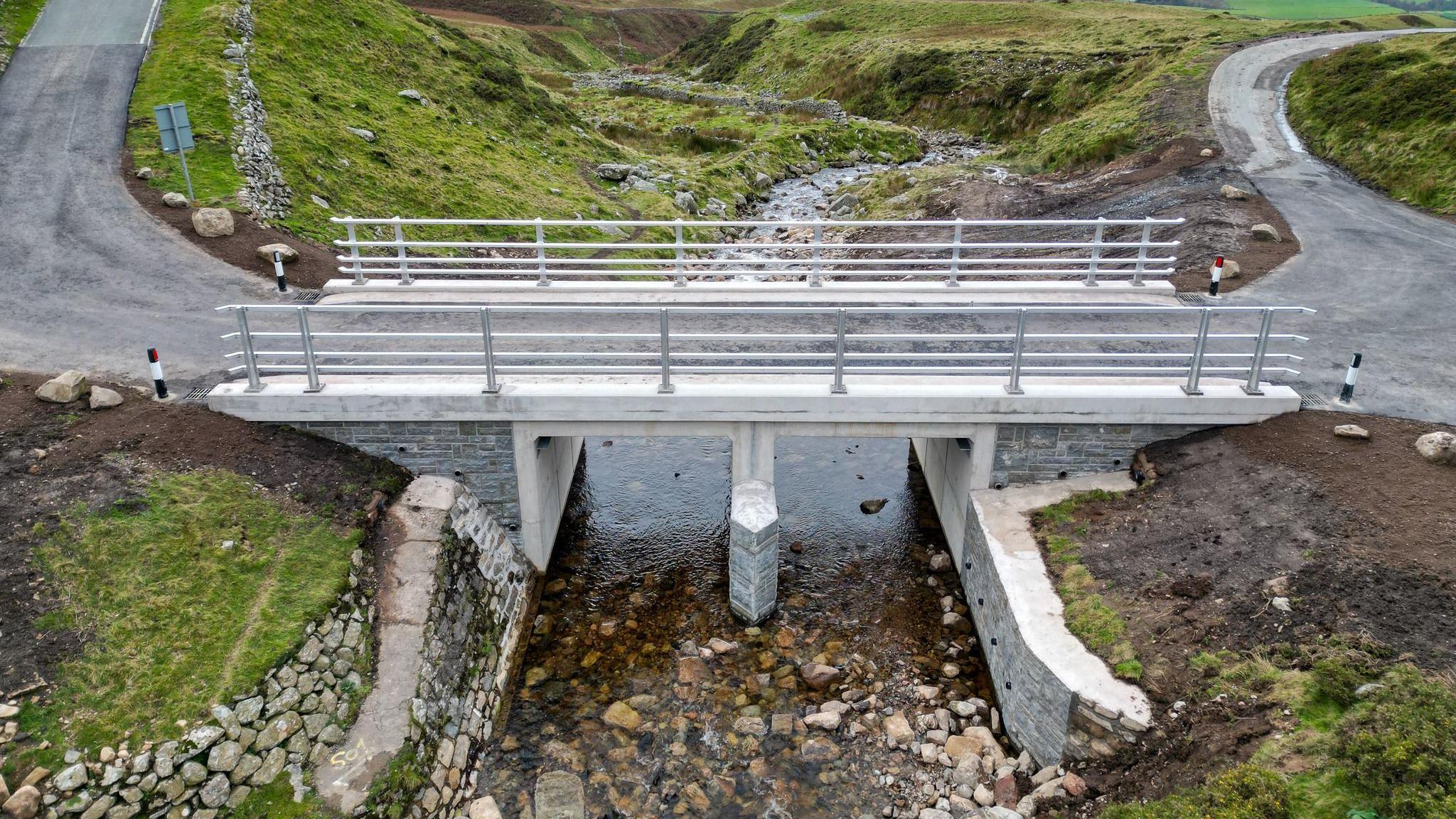The newly completed Buckbarrow Bridge. The bridge is made of concrete and stone brick and carries a single carriageway. It is entirely grey and has metal barriers on either side. A clear stream runs underneath the bridge.