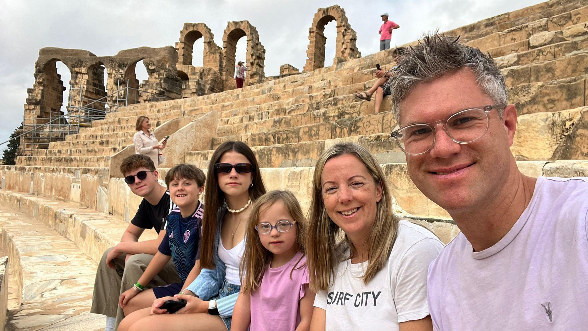 A family-of-six sit on the steps of an amphitheatre. The dad in the foreground has spiky grey hair and is wearing a purple T-shirt. Four children, two boys and two girls of different ages, sit in the background. The two older children are wearing sunglasses.