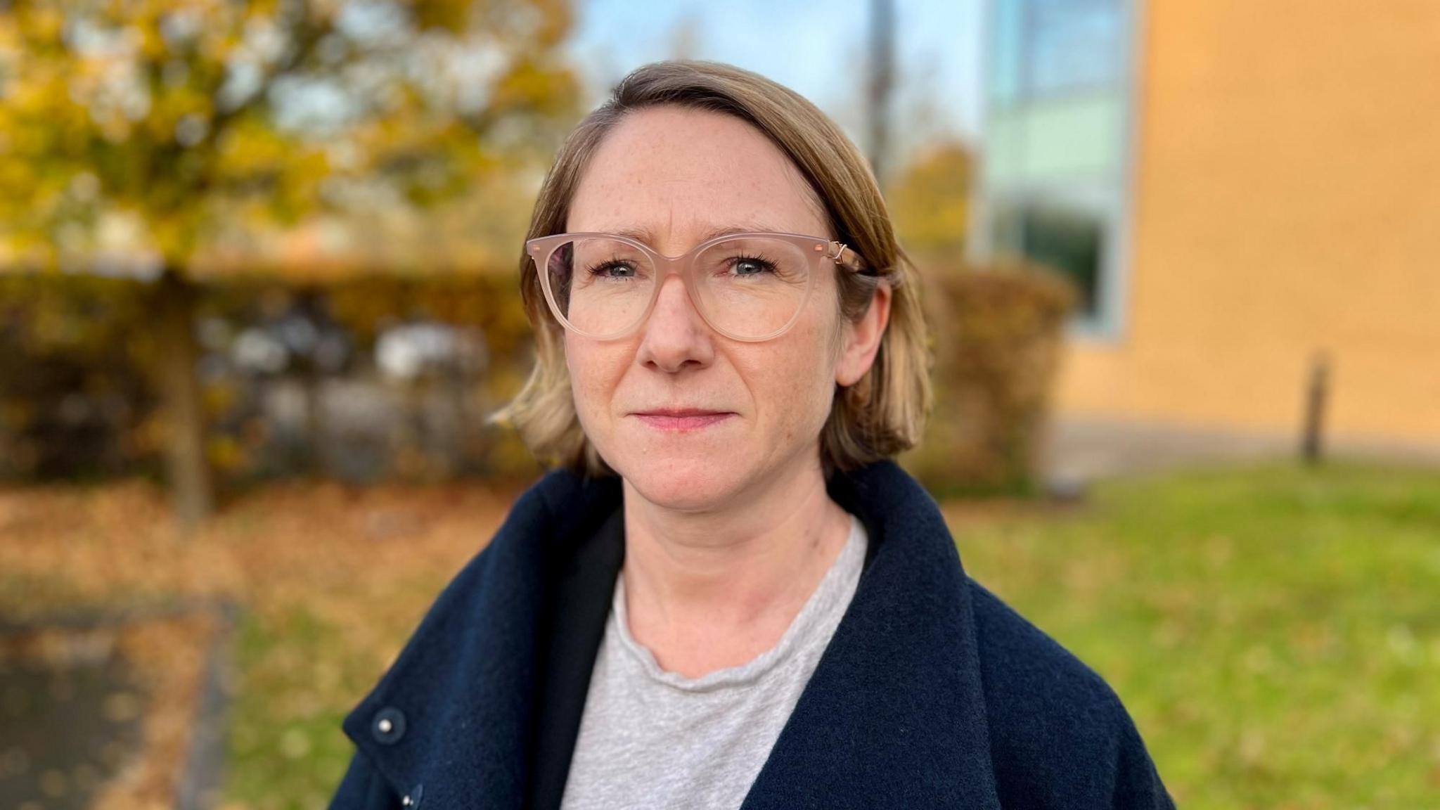 Saoirse Kerrigan is wearing a dark blue coat and a grey T-shirt. She has light pink coloured almost transparent glasses and has short bobbed mousey blonde hair. She is looking at the camera and is photographed outside. 