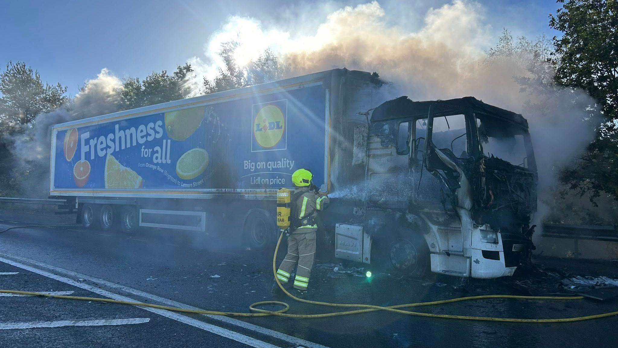 A Lidl lorry smoulders with a firefighter hosing it down by the roadside
