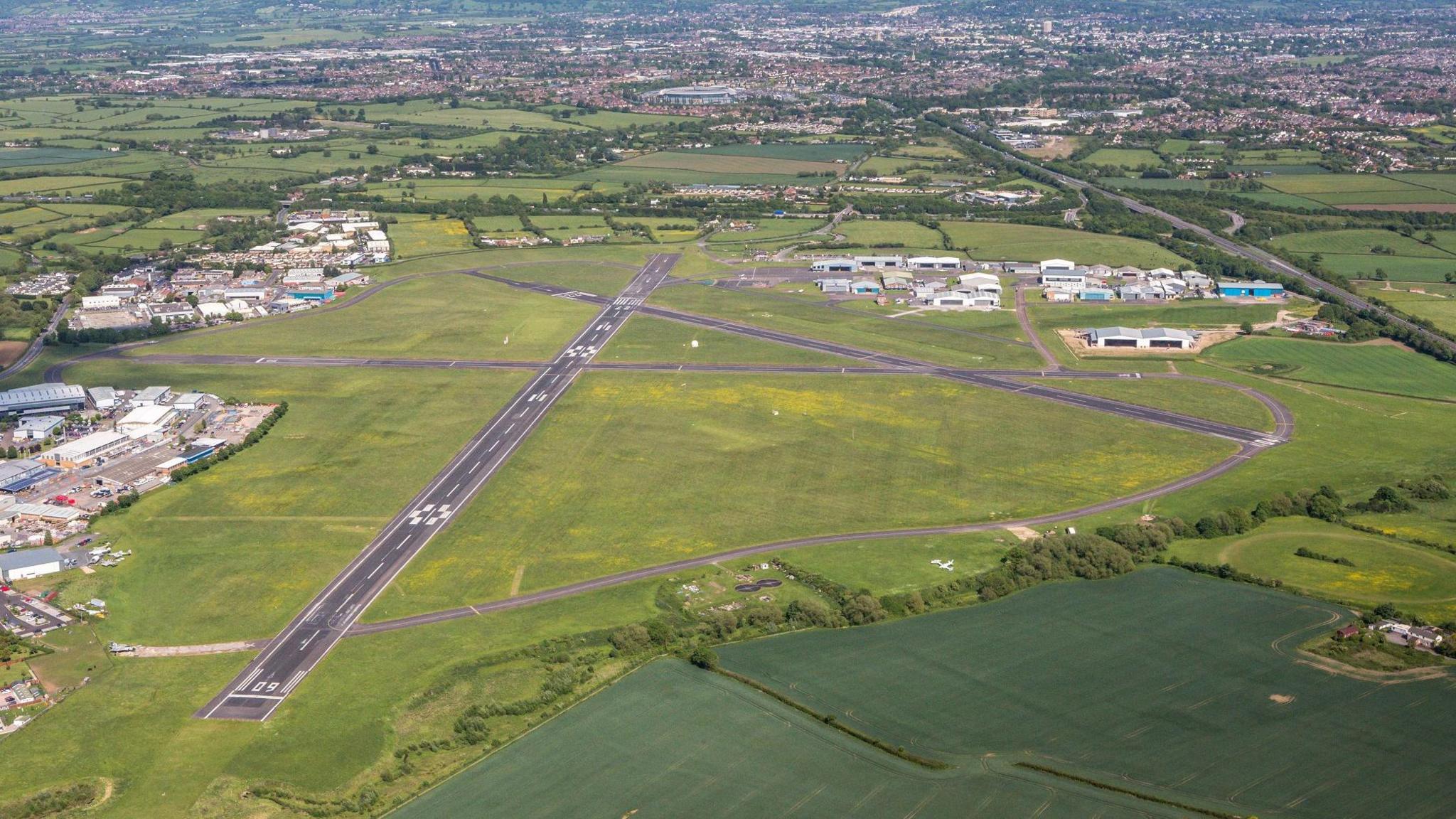 Aerial image of Staverton Airport - a small airport - in Gloucestershire. There are three runways that intersect one another on an airfield with hangars to the right. Cheltenham can be seen behind.