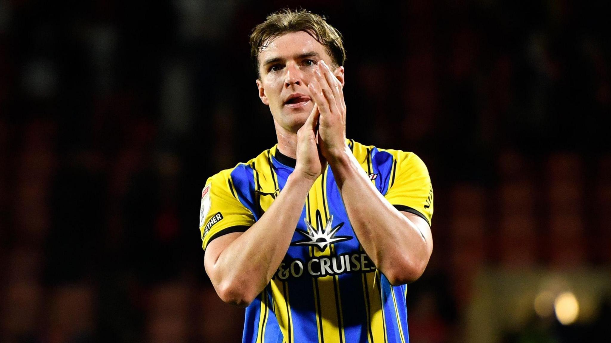 James Bree claps fans with hands to side of his face, wearing blue and yellow away kit