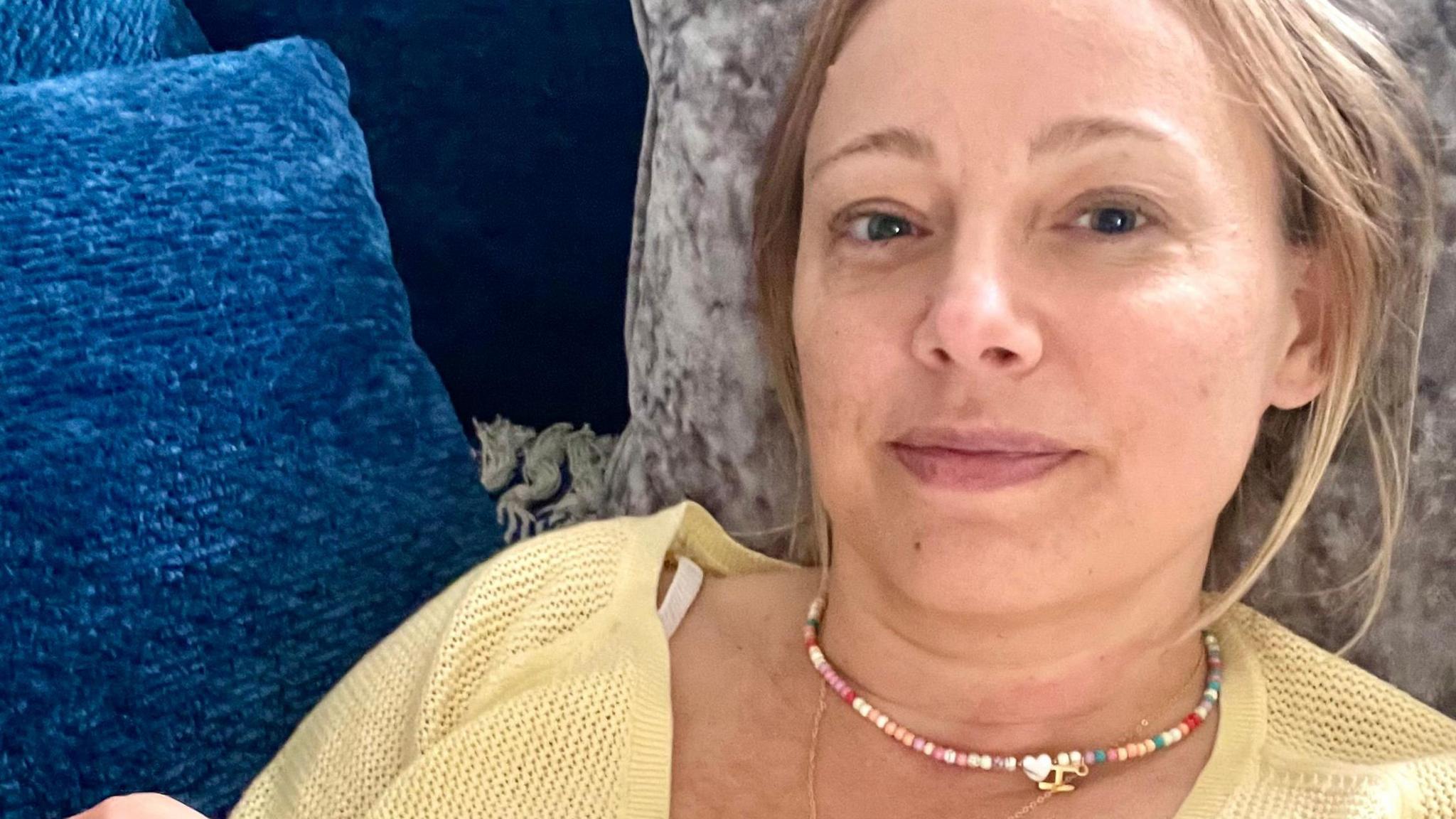 Hazel has long, blonde hair, is wearing a yellow cardigan and a colourful necklace, and is lying back on some blue and grey cushions