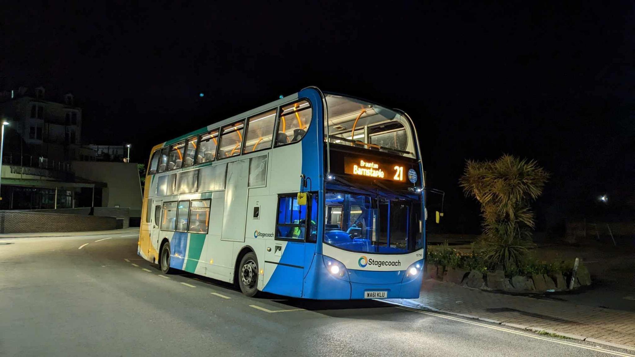 A picture of the night bus parked up at a bus stop. It is a mainly white bus with a blue front and a multi coloured end section.