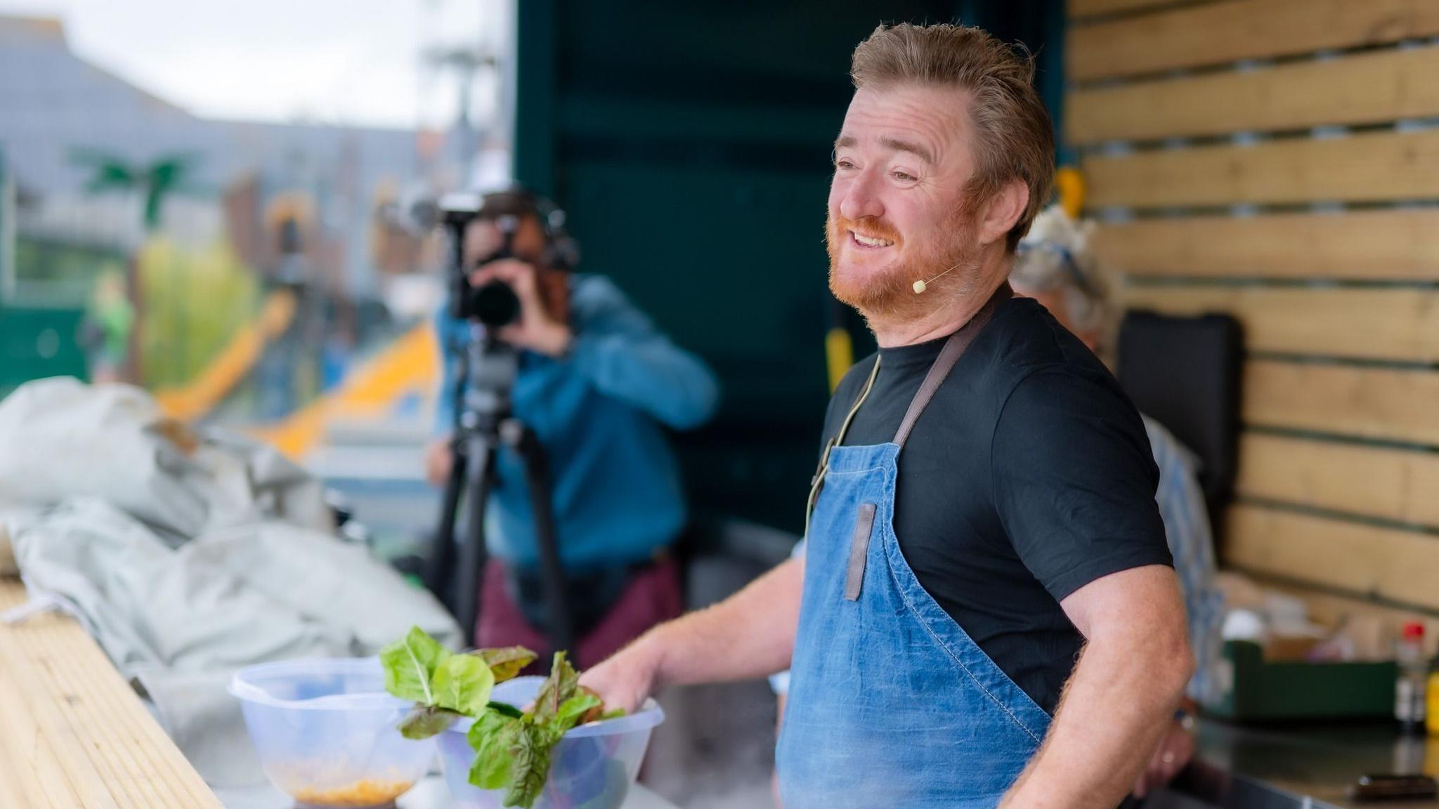 Jack Stein gives a cooking demonstration. He has fair hair and a ginger beard, and is wearing a black t-shirt and denim apron. A television cameraman, standing to the left, is filming the event.