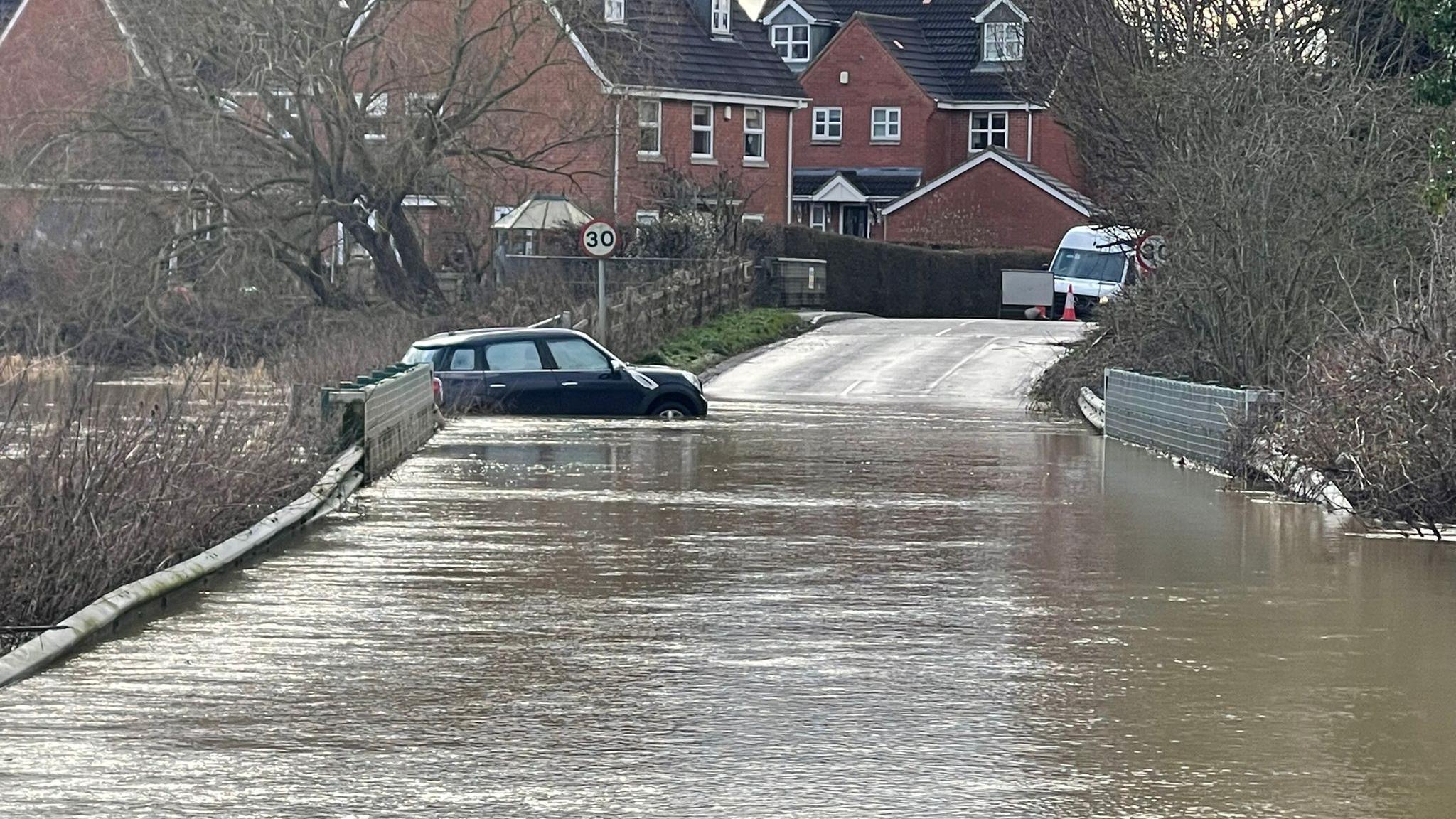 A bridge leading up to a residential street is inundated with brown flood water. A black Mini car is stuck next to the bridge