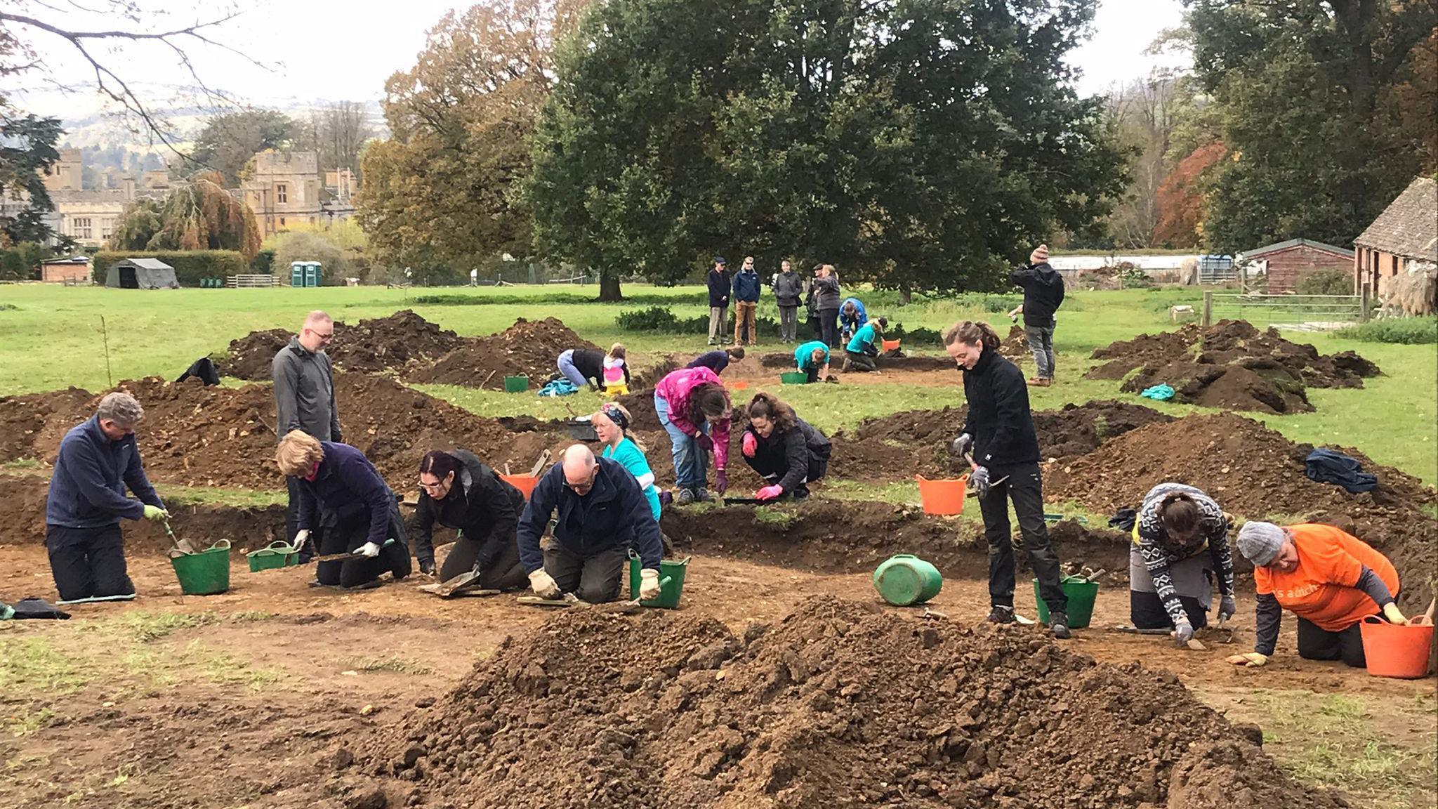 People in archaeological trenches, digging on their hands and knees to uncover a medieval village. Mounds of soil surround them as they use trowels to scoop it out into flexible buckets. The dig is taking place on the grounds of a castle, which can be seen lurking behind trees with browning leaves in the background.