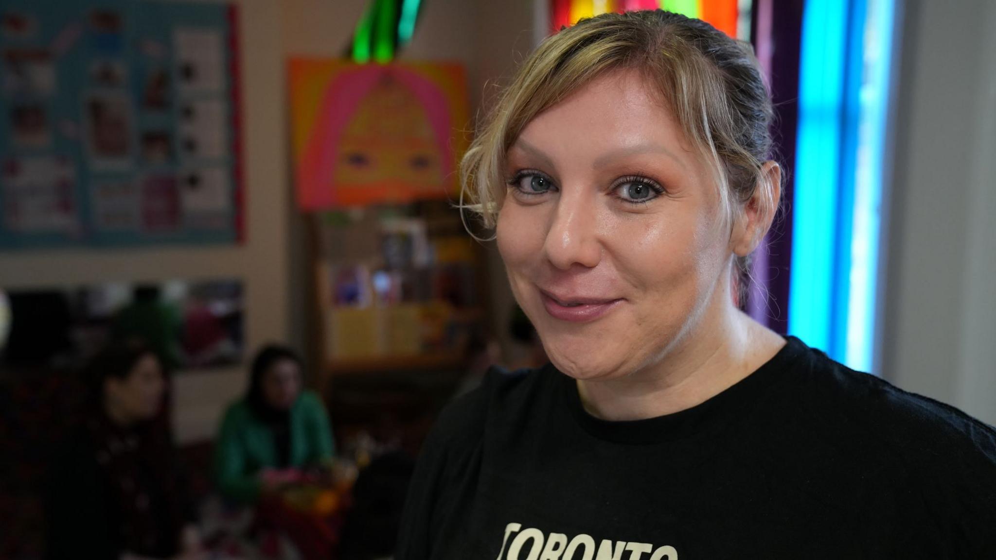 Hannah Aria has blonde hair and wearing a black top, she is smiling at the camera. In the background a few women are sat together talking. 