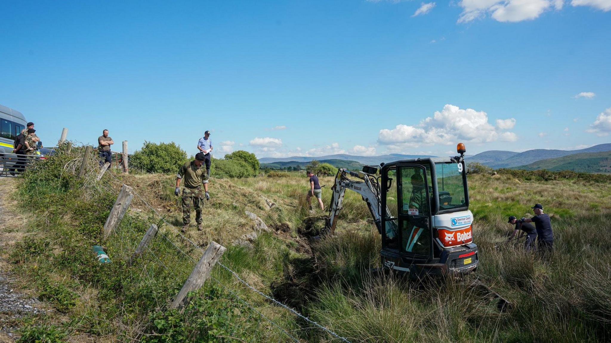 A red and grey mini digger is removing pieces of grass from the ground. Around the digger a collection of people are watching, some are wearing camouflage clothing while others are dressed in navy. The land is cordoned off by a wire fence. At the edge of the land a vehicle is sitting. In the background is a row of mountains.