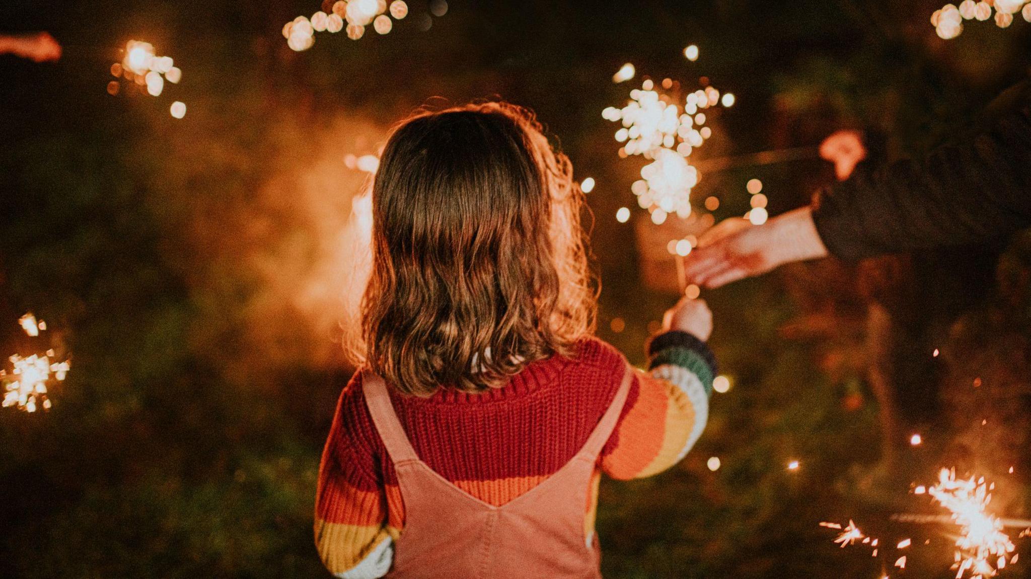 Child is seen holding a sparkler with an adult's hand also in shot helping her keep it upright. She is facing away from the camera standing on a grass surface.