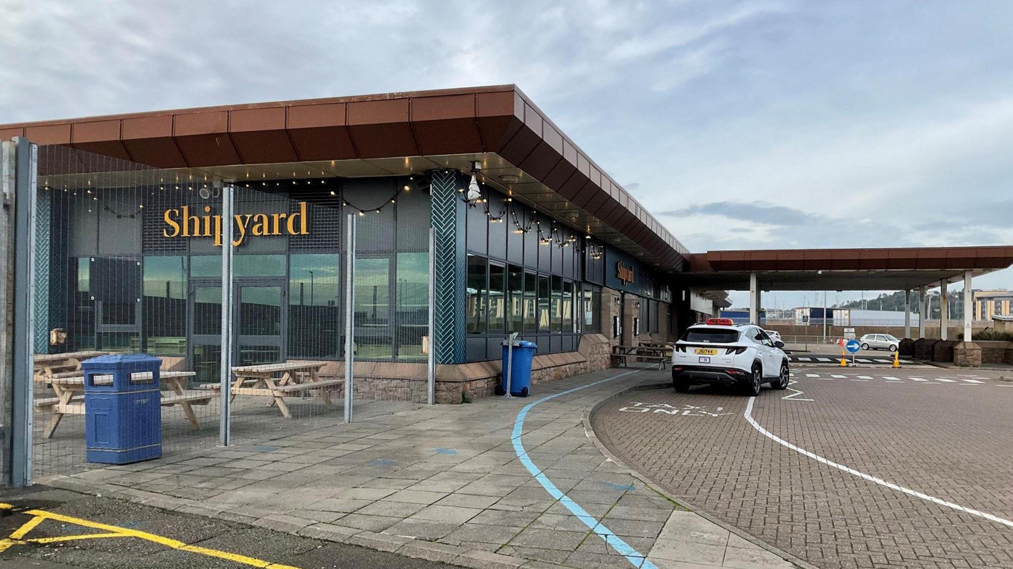 An outside photo of the Shipyard cafe at Elizabeth Harbour in Jersey. There is a white car parked outside. It's a large building with long glass windows. The word shipyard is prominent in yellow writing on the wall. There are benches and a bin behind a metal fence.