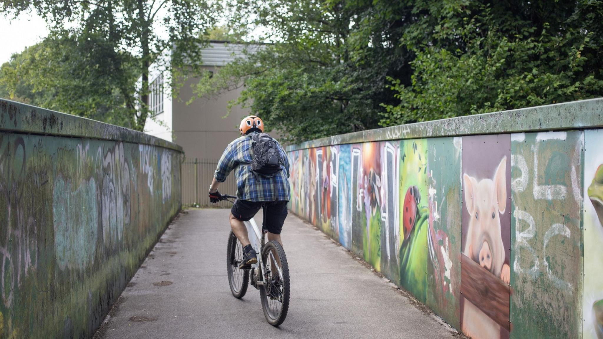 A cyclist in a checked shirt and shorts travelling over a bridge in Hengoed, Caerphilly county, which has graffiti on the walls
