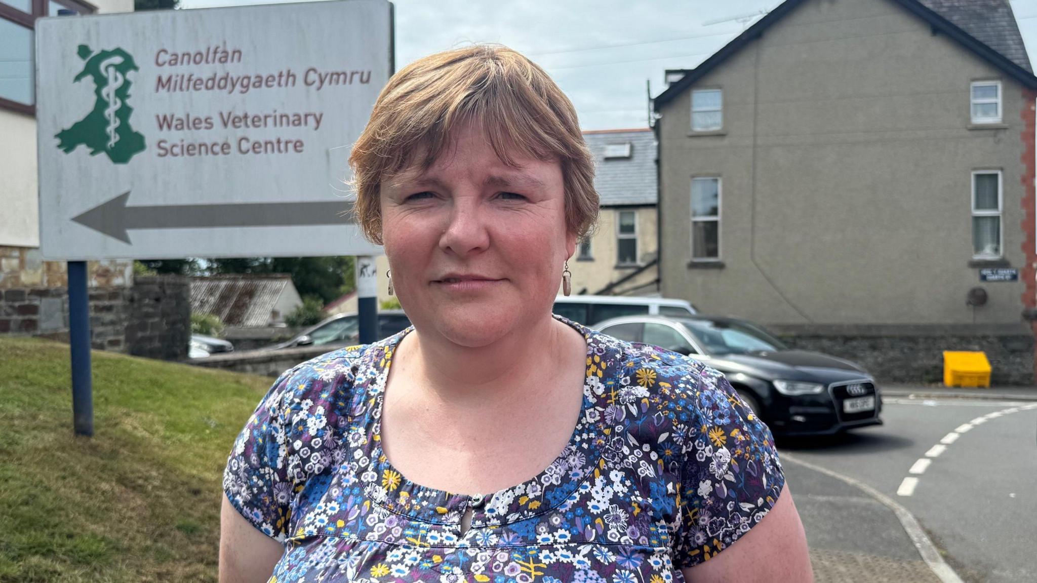 Mae'r milfeddyg yn sefyll o flaen y arwydd Canolfan Milfeddygaeth Cymru mewn top blodeuog. 