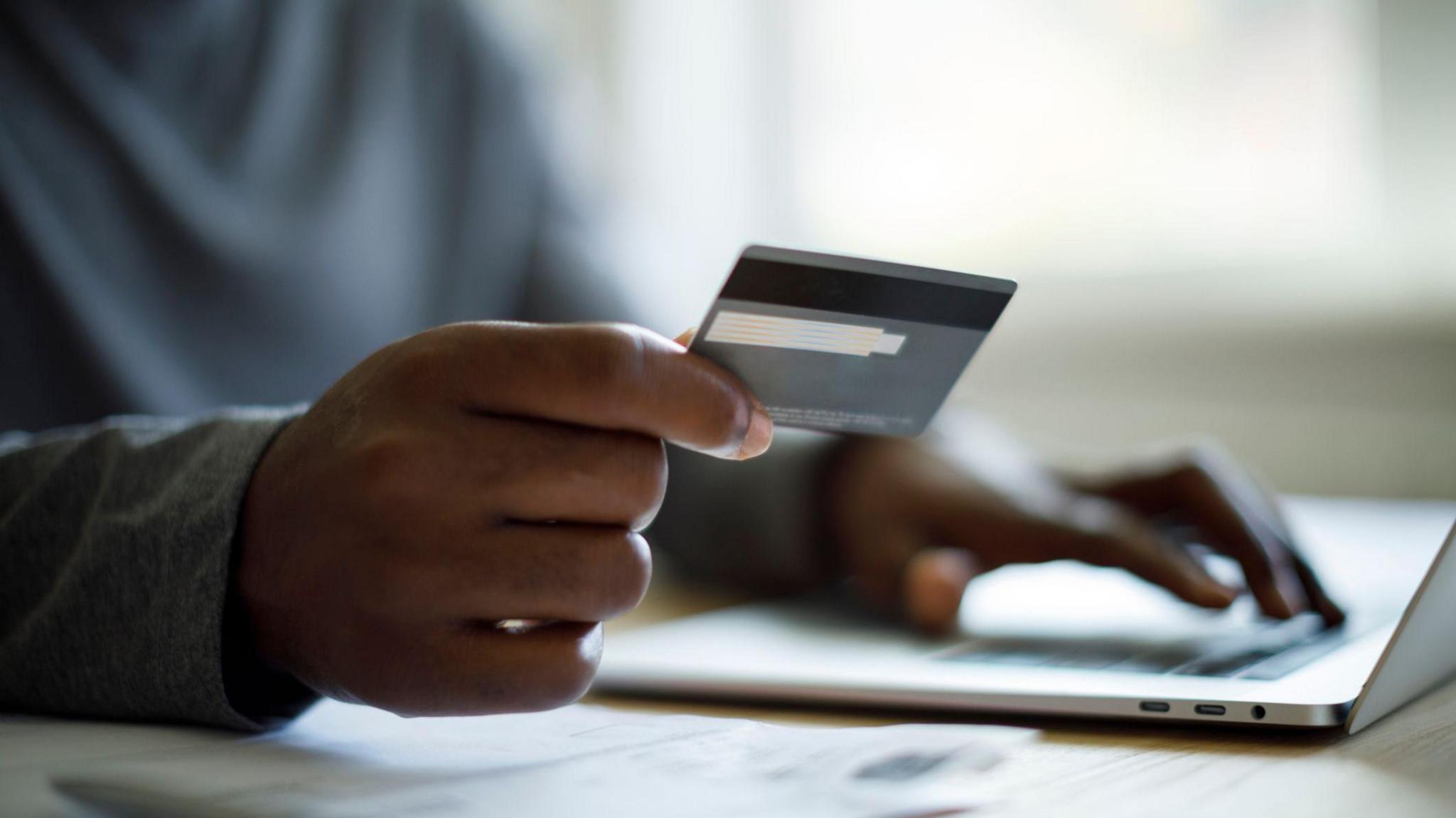 A man wearing a grey sweater sitting at a desk in front of a laptop holds a credit card in one hand between his index finger and his thumb, while his other hand is tapping the keys on the laptop. We cannot see the face of the man.