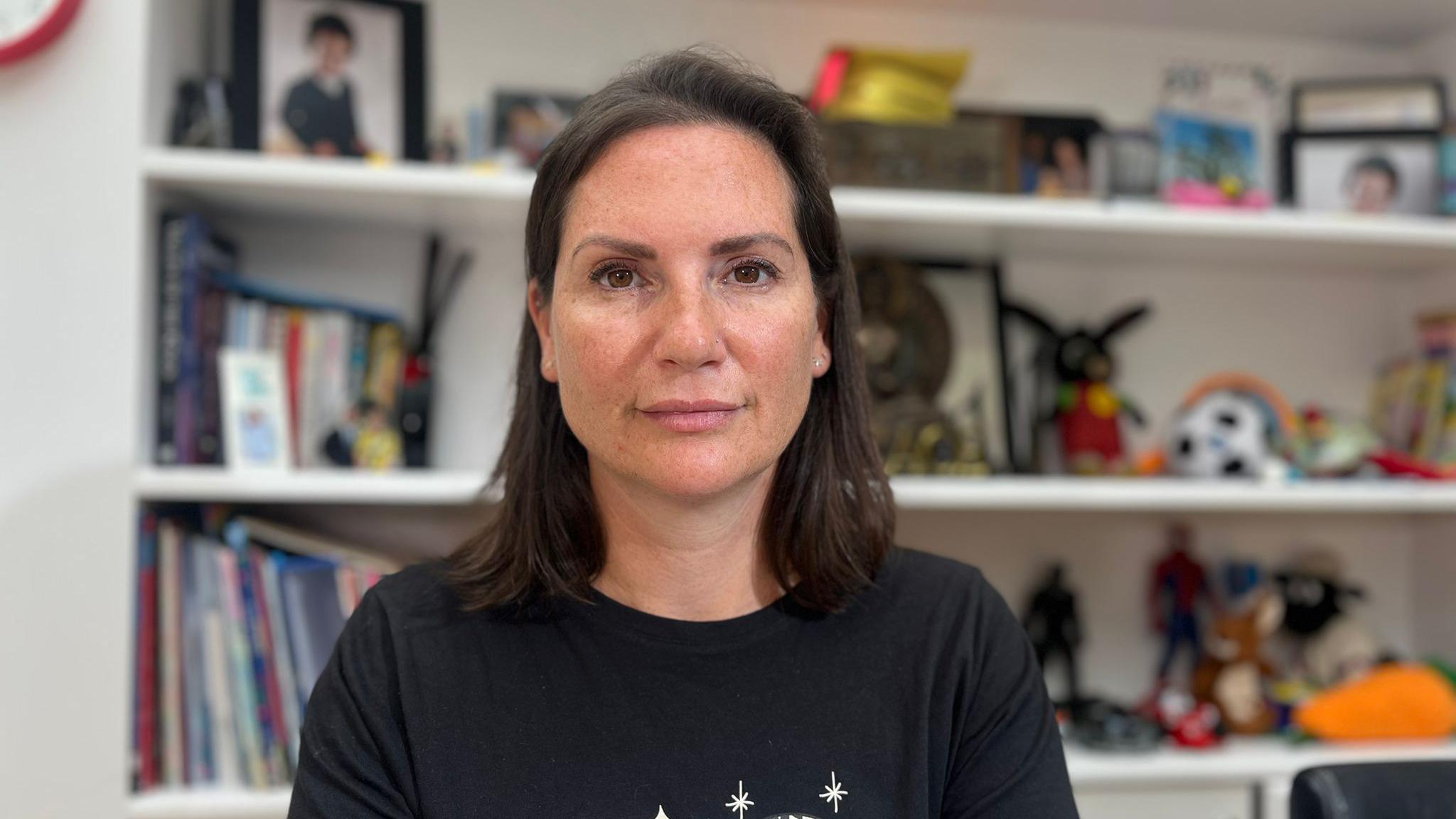Natalie Neri looks into camera with photos of her son, books, and toys on the shelf in the background. She has shoulder-length dark hair and is wearing a black t-shirt with white stars on